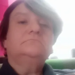 Portret kobiety w szarej bluzie z ciemnym kołnierzykiem, z kr&oacute;tkimi ciemnymi włosami z grzywką, na tle rozmytej r&oacute;żowej ściany i jasnego okna.