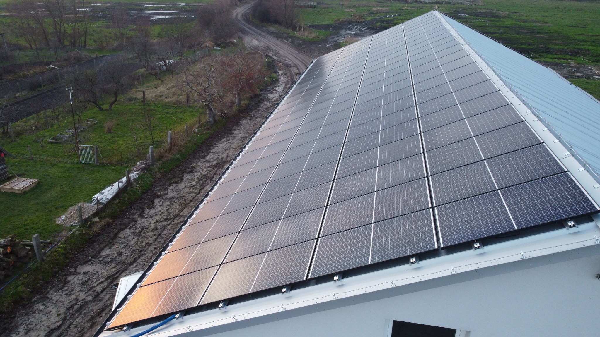 Instalacja fotowoltaiczna na dachu budynku jednorodzinnego, widok z góry, z widoczną polną drogą i zielonym terenem w tle.