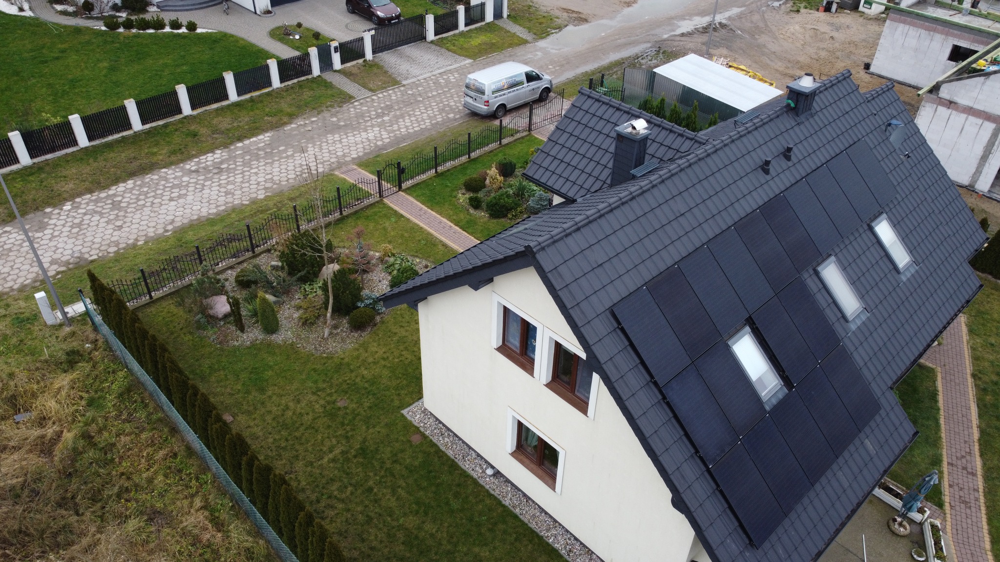 Widok z góry na dom jednorodzinny z panelami fotowoltaicznymi na dachu, regularne ułożenie paneli, dachówka ceramiczna, okna dachowe, zadbany trawnik i ogród, na podjeździe zaparkowany biały bus.