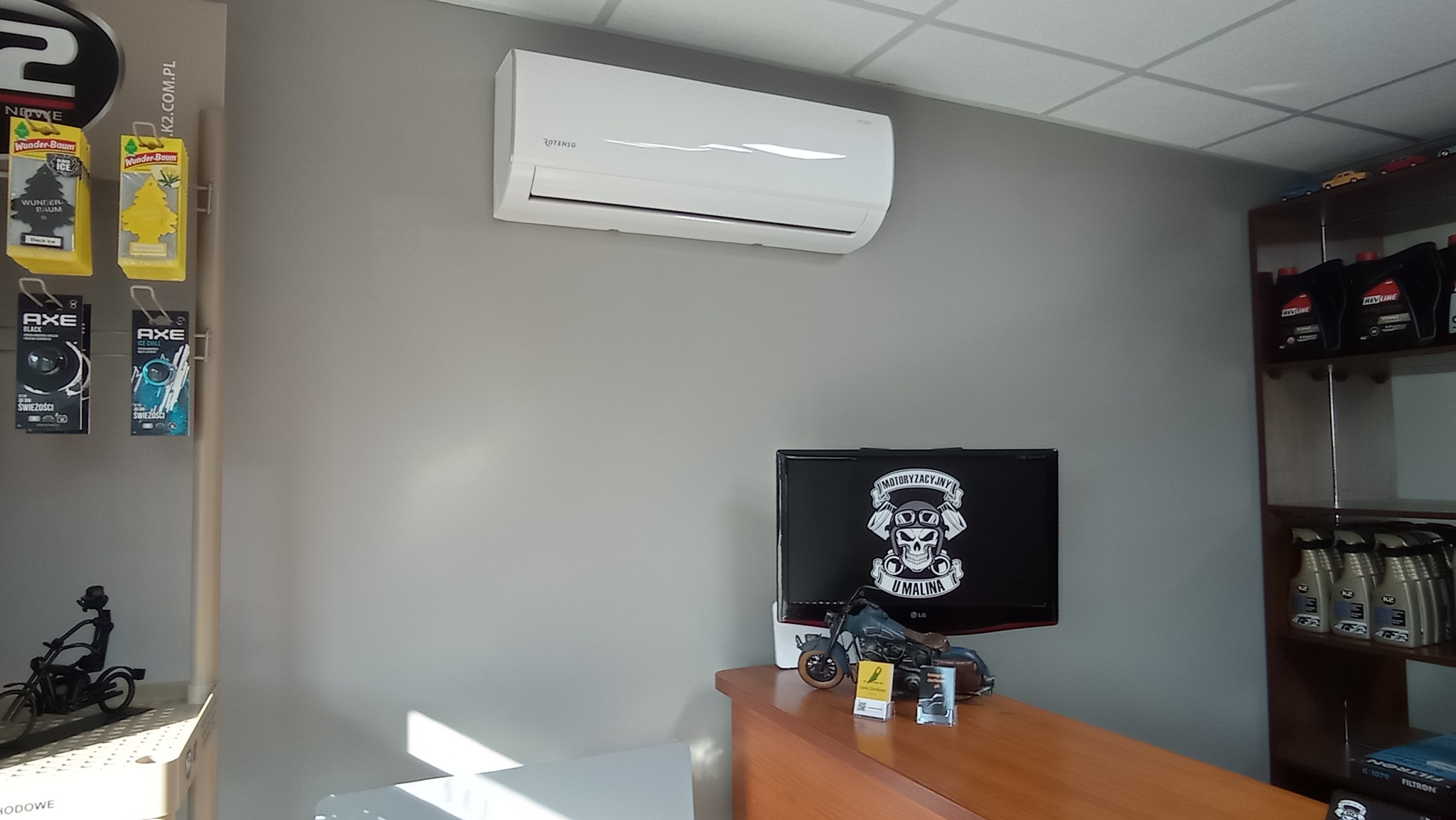 Biały klimatyzator Rotenso zamontowany na szarej ścianie w pomieszczeniu, widoczne dekoracje motoryzacyjne i zapachy samochodowe.