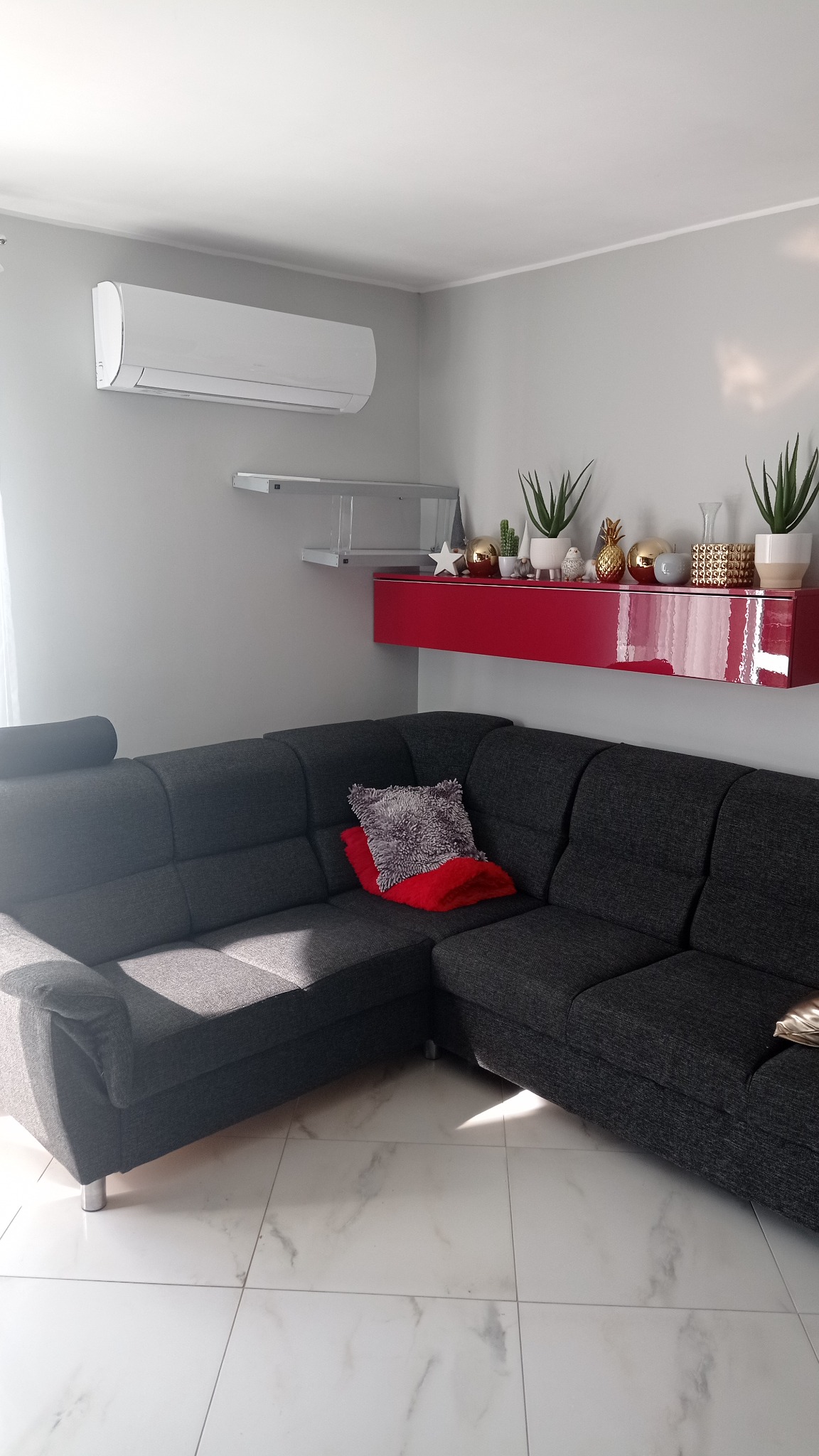Biały klimatyzator ścienny w nowoczesnym salonie z szarą sofą narożną, czerwonym regałem i marmurową podłogą.