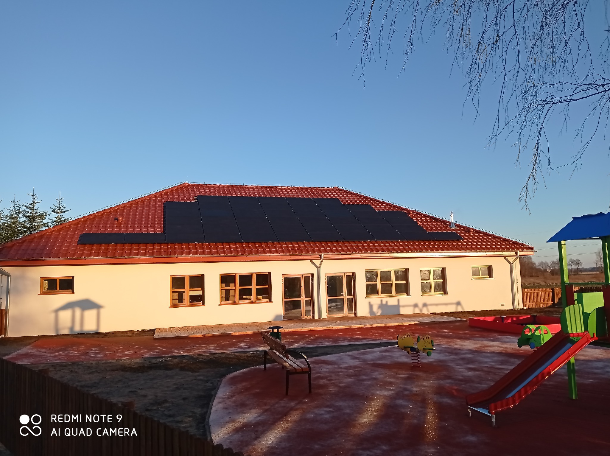Budynek przedszkola z panelami słonecznymi na czerwonym dachu, plac zabaw z huśtawkami i zjeżdżalnią na pierwszym planie, widok w słoneczny poranek.