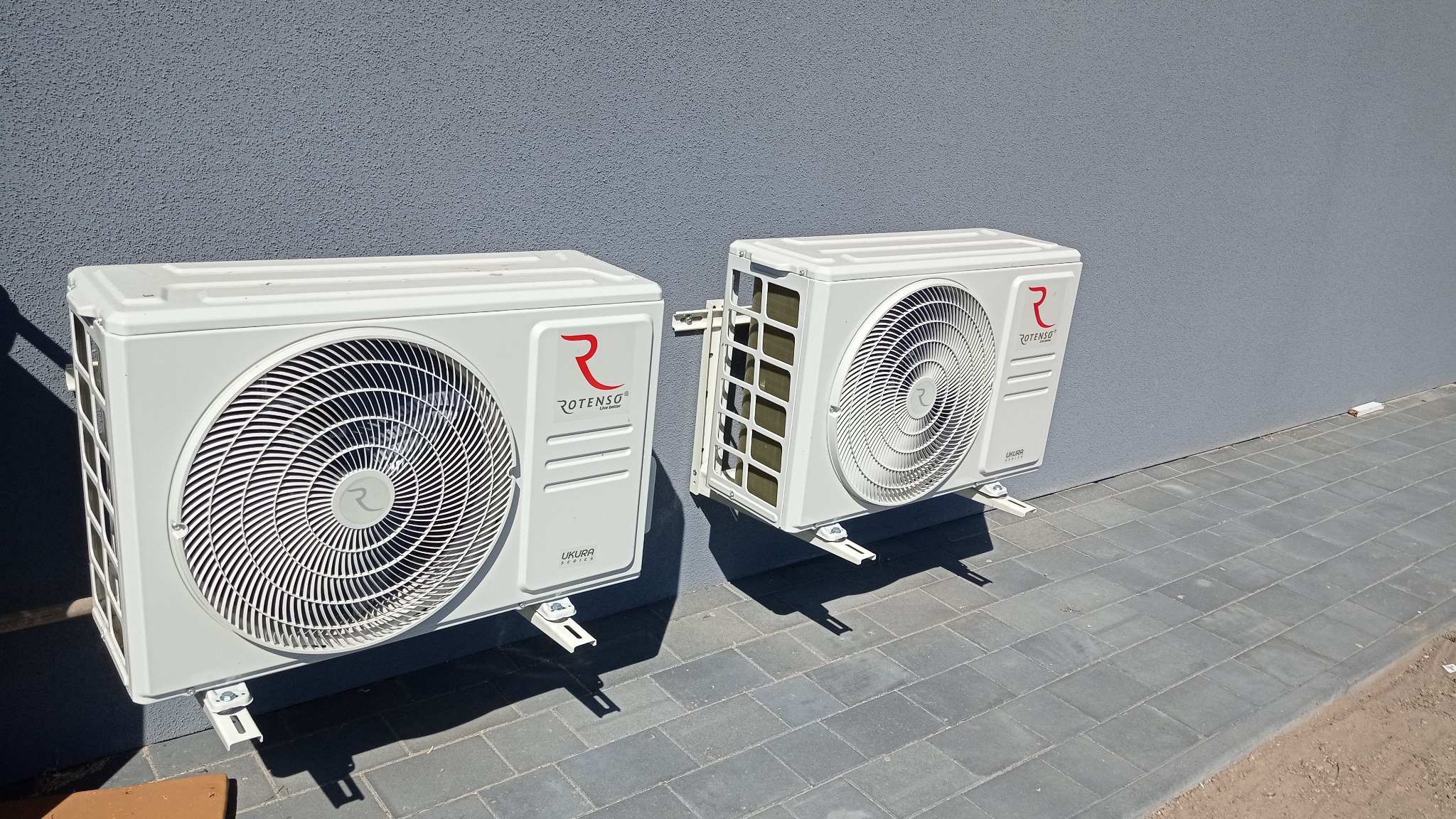 Dwa białe klimatyzatory marki Rotenso zamontowane na szarej elewacji budynku, widoczne przewody i wsporniki montażowe, na pierwszym planie szara kostka brukowa.
