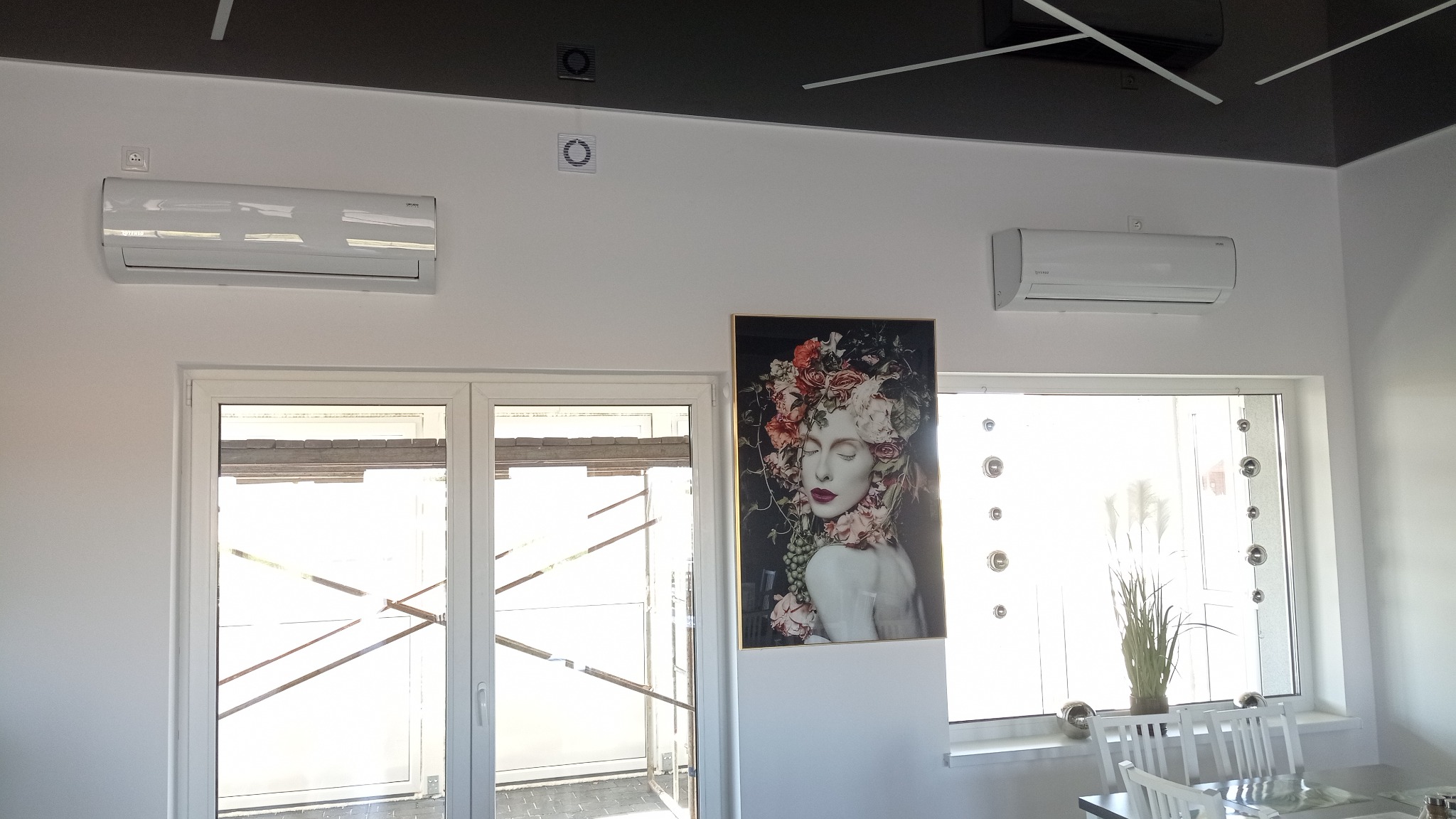 Dwa białe klimatyzatory zamontowane na ścianie nad oknami, pomiędzy nimi wisi oprawiony obraz z motywem kwiatowym, widoczne kratki wentylacyjne na suficie.
