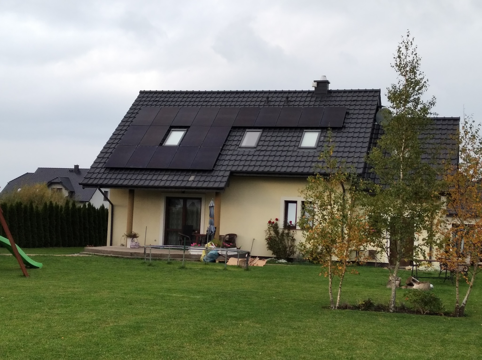 Dom jednorodzinny z ciemną dachówką i panelami słonecznymi wkomponowanymi w połać dachu, widok z ogrodu w pochmurny dzień.