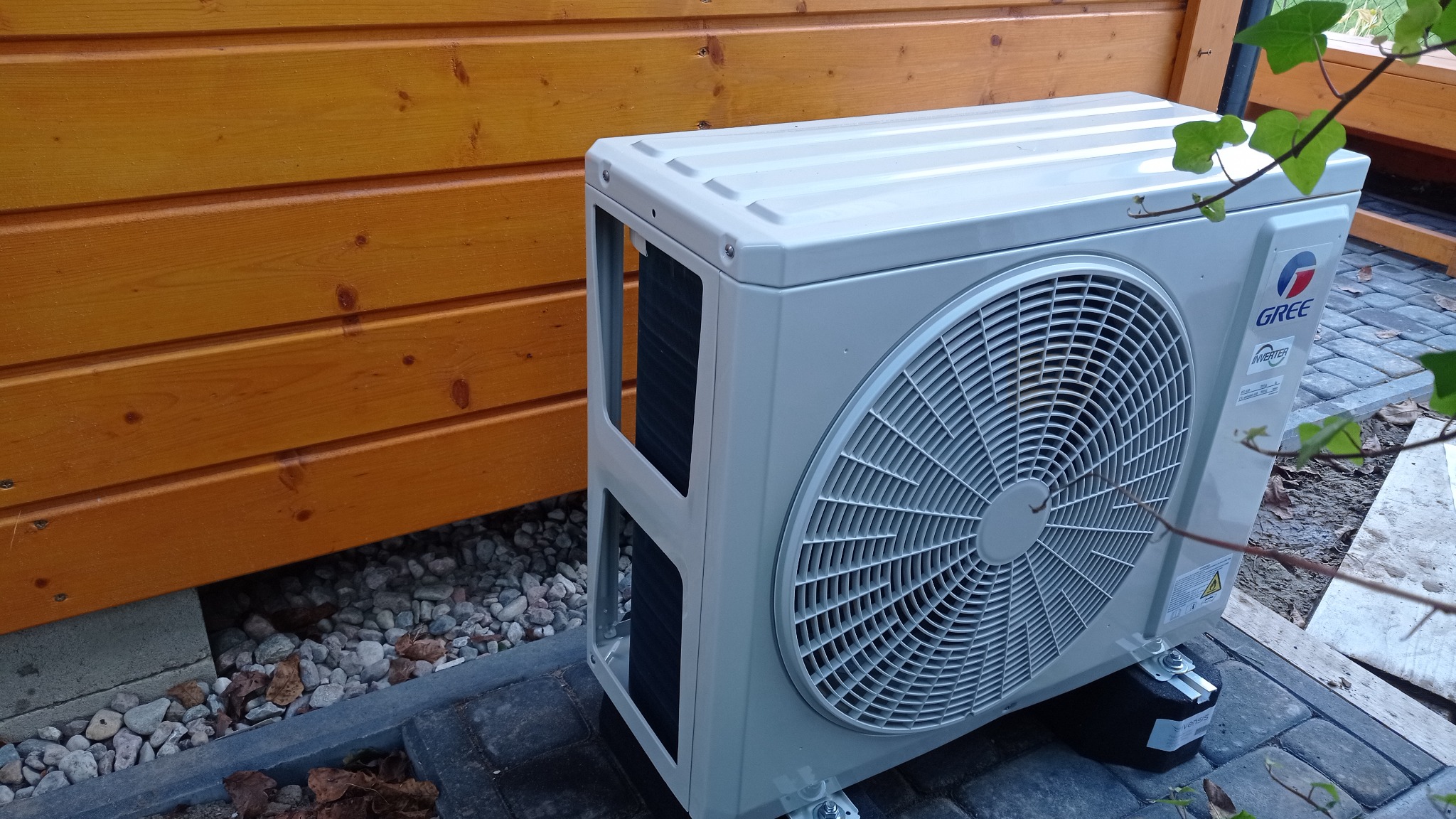 Zewnętrzna jednostka klimatyzacji GREE zamontowana na kostce brukowej przy drewnianej elewacji budynku, z widocznymi elementami instalacji i fragmentami roślinności.