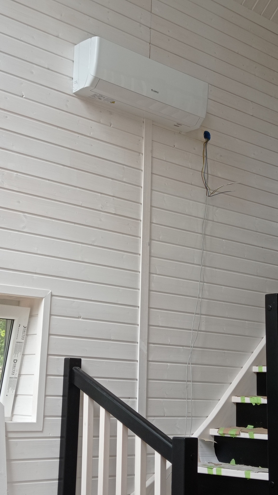 Biały klimatyzator Gree zamontowany na ścianie z białych paneli boazeryjnych w domu, widoczne wystające kable, fragment schodów z czarną balustradą i zabezpieczone taśmą stopnie.