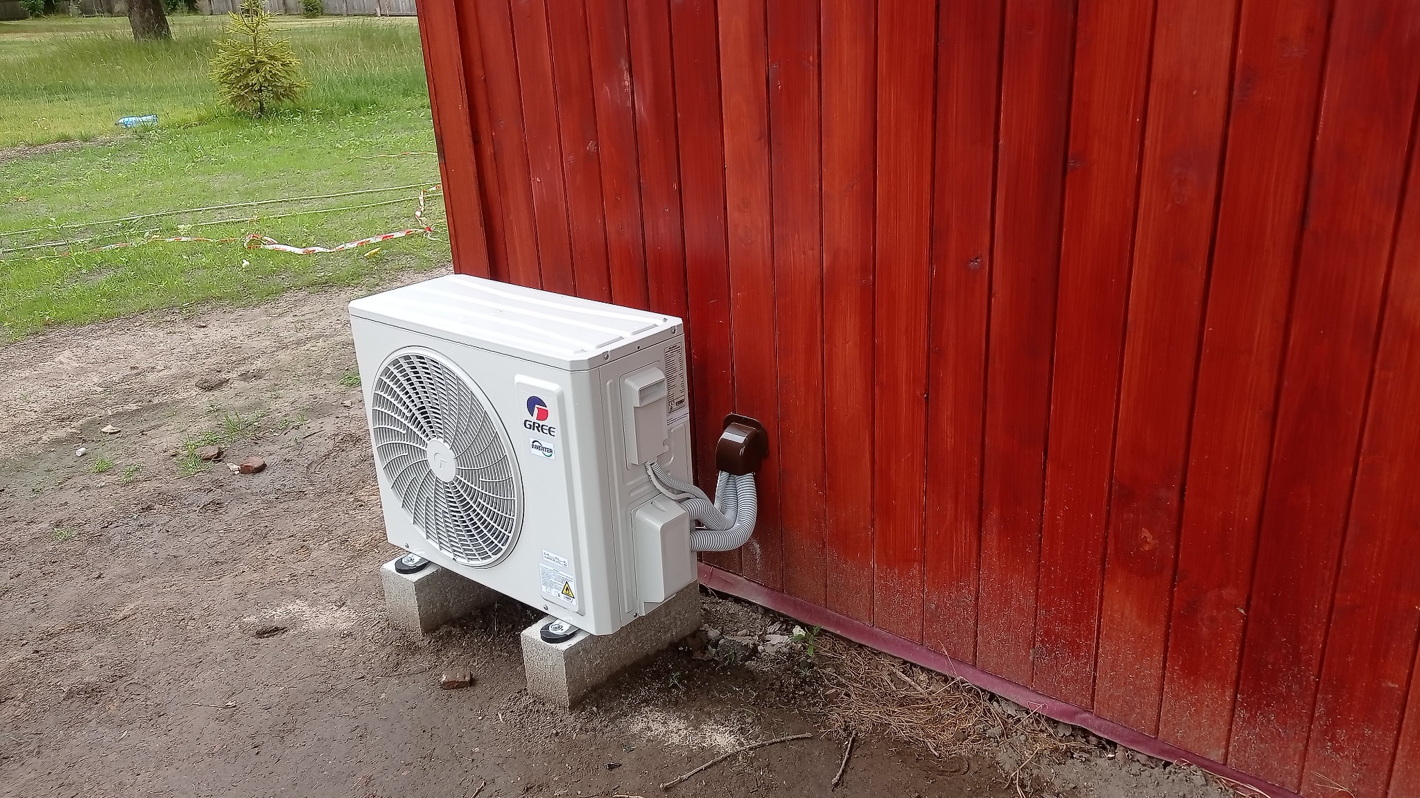 Zewnętrzna jednostka klimatyzacji marki Gree zamontowana na betonowych bloczkach przy czerwonej, drewnianej ścianie budynku, widoczne przewody prowadzące do ściany.