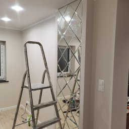 KAL-BUD Kamil Kalbarczyk - Montaż dekoracyjnego panelu lustrzanego na ścianie w trakcie prac wykończeniowych, widoczna drabina, narzędzia i fragment podłogi.