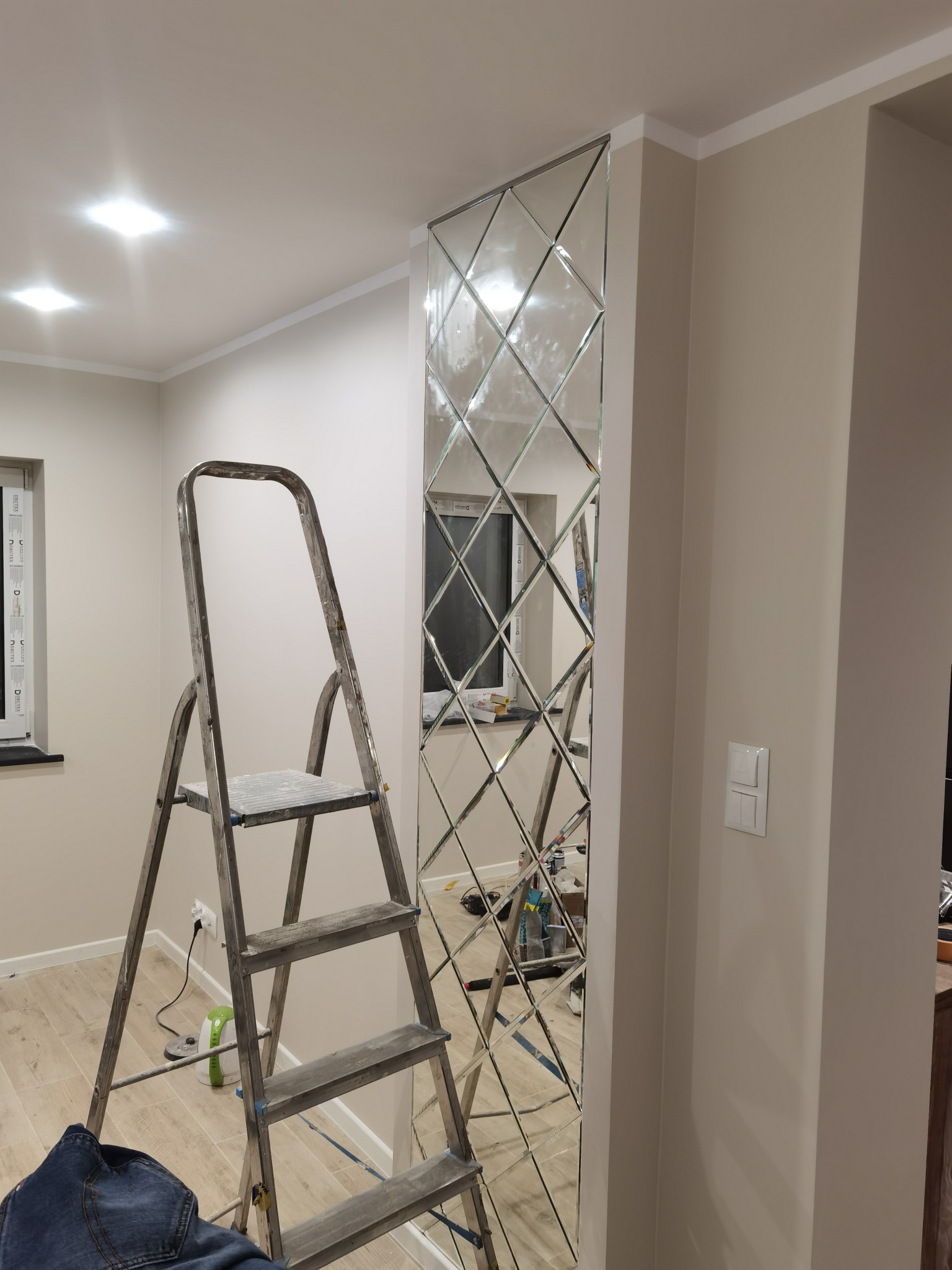 Montaż dekoracyjnego panelu lustrzanego na ścianie w trakcie prac wykończeniowych, widoczna drabina, narzędzia i fragment podłogi.