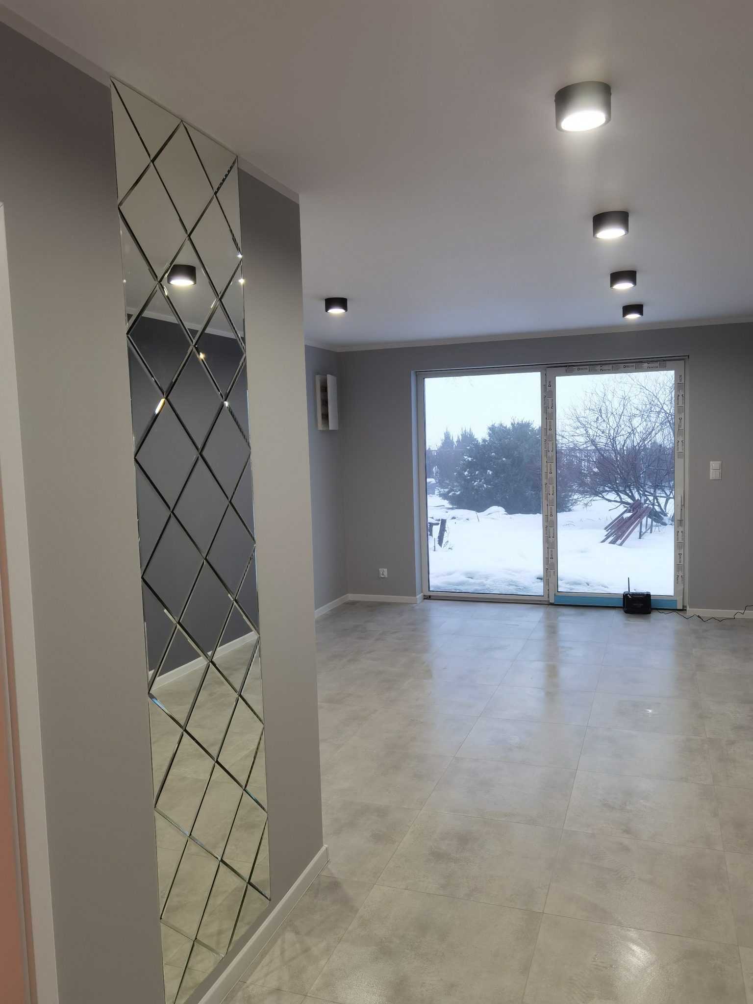 Szare wnętrze po odświeżeniu, z pionowym lustrem w romby na ścianie, szarą podłogą z płytek i oknem balkonowym z widokiem na zaśnieżony ogród.