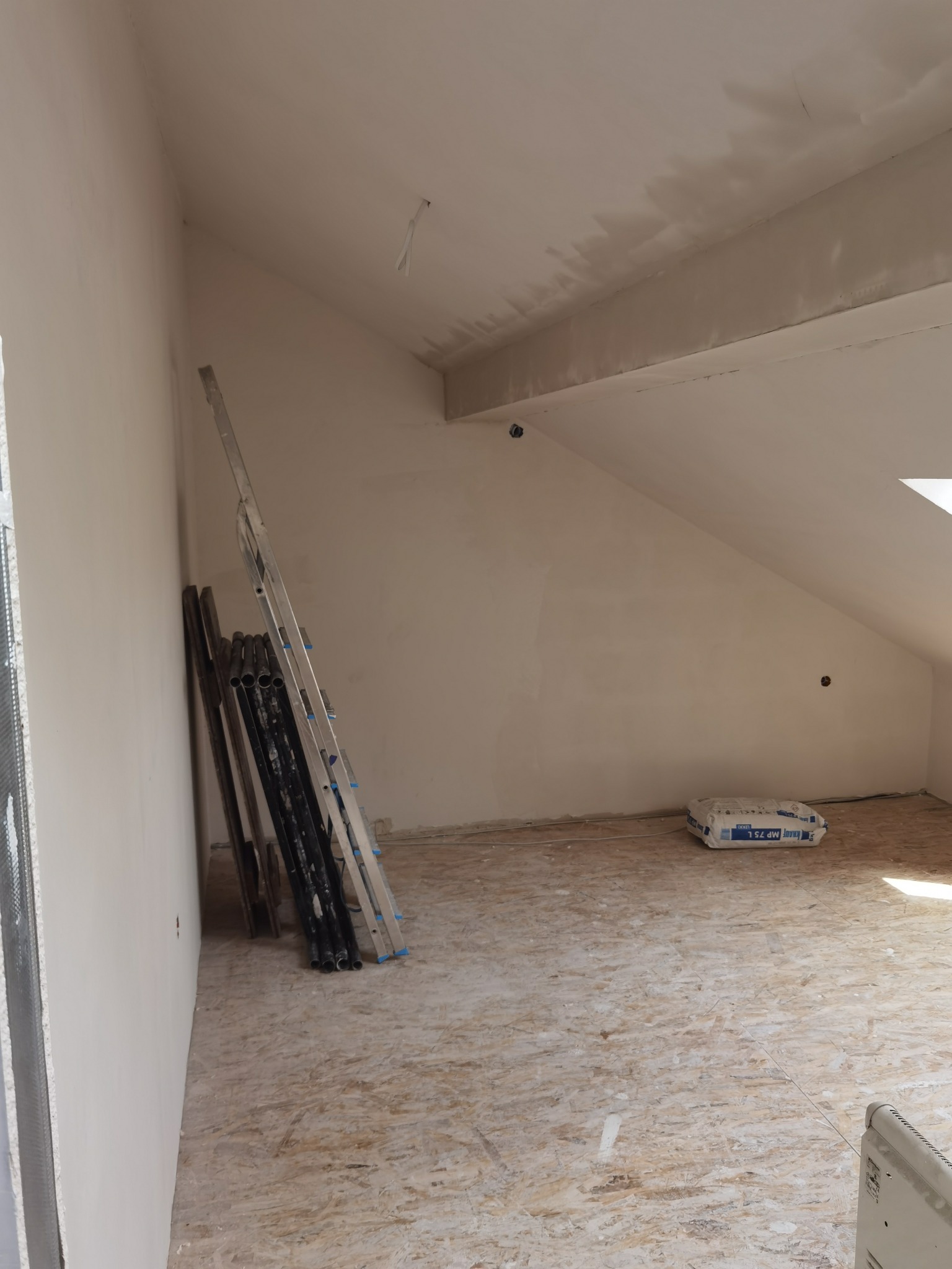Poddasze w trakcie prac wykończeniowych: ściany po gładziowaniu, widoczna drabina, rury i worek gładzi Knauf MP 75 L na podłodze z płyt OSB.