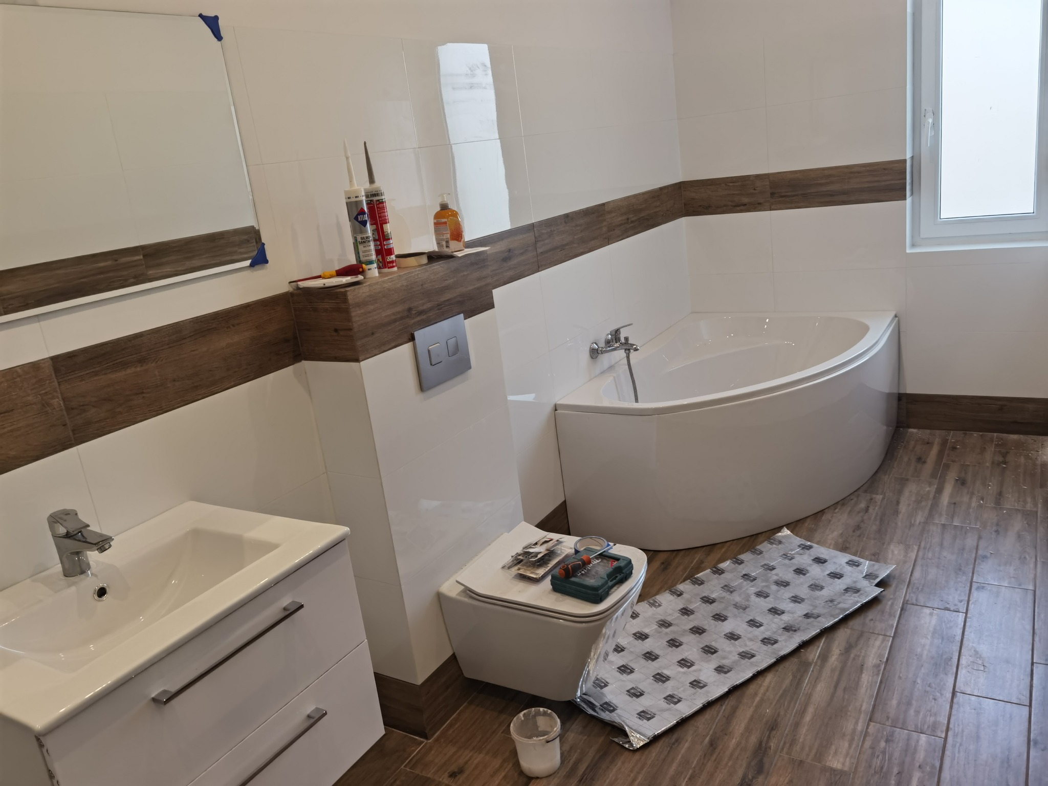 Biała łazienka z wanną narożną, umywalką z szafką i toaletą w trakcie montażu, widoczne narzędzia i materiały budowlane na desce sedesowej, płytki imitujące drewno na podłodze i pasie na ścianie.