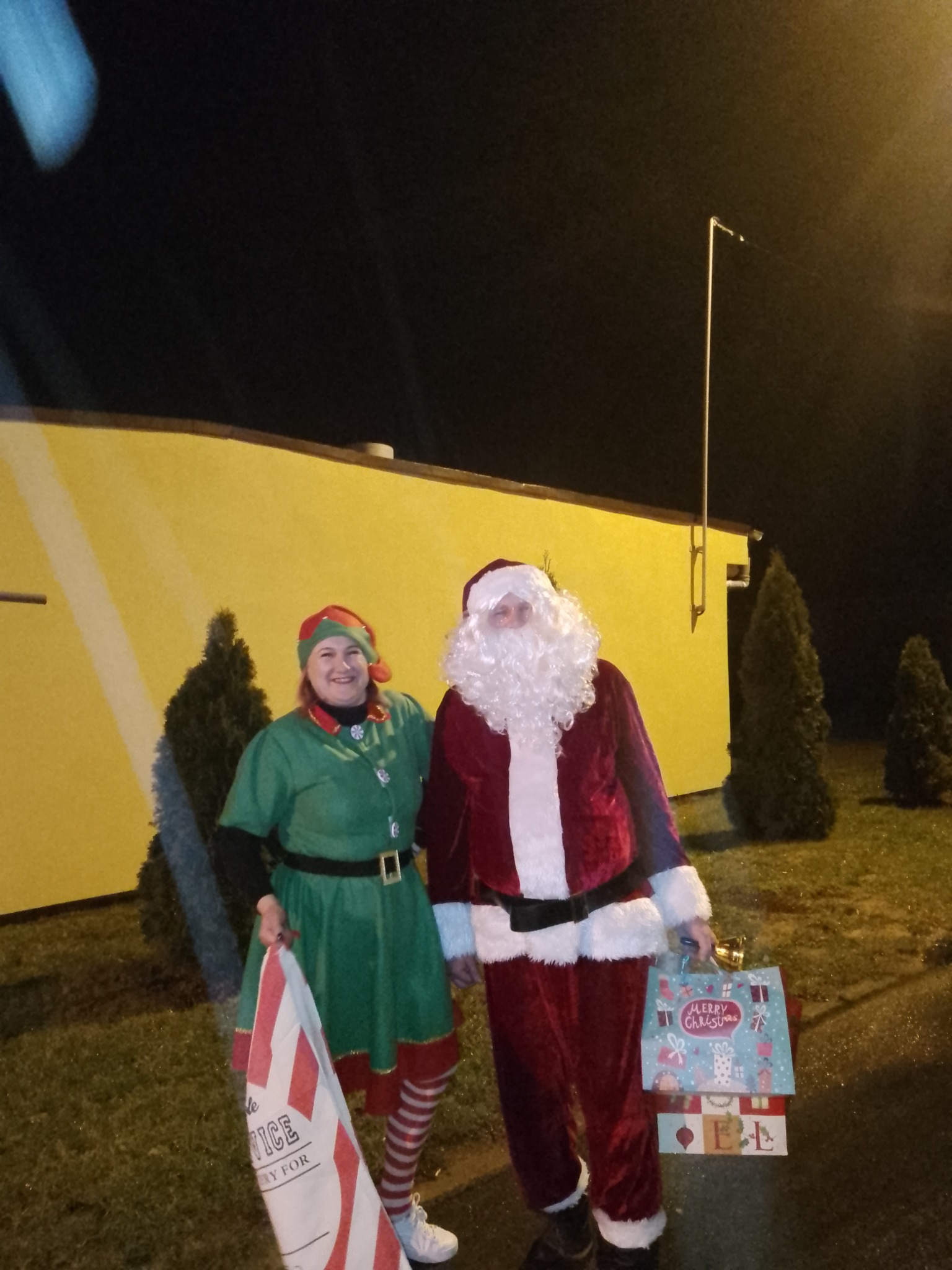 Kobieta przebrana za elfa i mężczyzna w stroju Świętego Mikołaja stoją przed żółtym budynkiem z torbami prezentowymi.