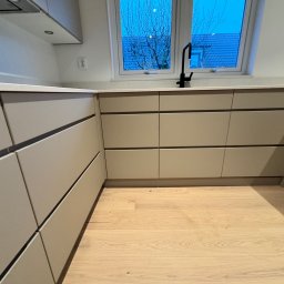 Renobuil - Nowoczesna, beżowa zabudowa kuchenna z drewnianą podłogą, czarną armaturą i widokiem na ogród przez okno.