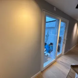 Świeżo pomalowane ściany w odcieniach szarości, widoczne przesuwne drzwi balkonowe z widokiem na podw&oacute;rze z narzędziami ogrodniczymi i materiałami budowlanymi, parkiet na podłodze, lampy sufitowe...