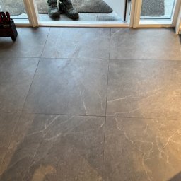 Renobuil - Szare, kwadratowe płytki podłogowe z marmurkowym wzorem, widoczne buty robocze przy progu i pojemnik z narzędziami w rogu pomieszczenia.