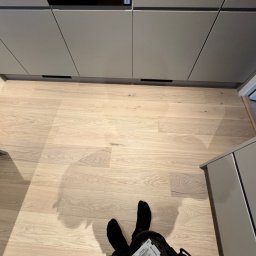 Renobuil - Jasny parkiet ułożony w jodełkę francuską, widok z góry na stopy w czarnych skarpetach i fragment szafek kuchennych w kolorze szarym.