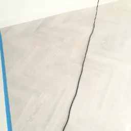 Jasny parkiet ułożony we wzór jodełki, zabezpieczony niebieską taśmą malarską przy ścianie, widoczny kabel zasilający wpięty do gniazdka.