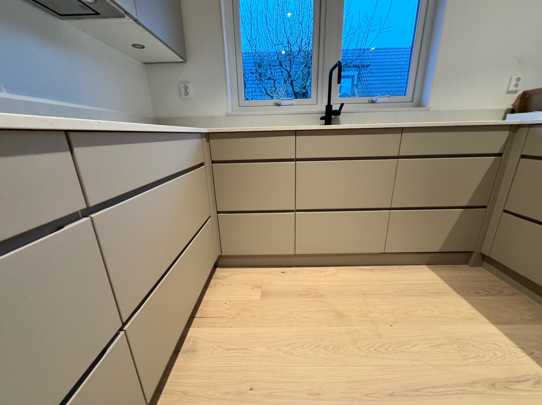 Nowoczesna, beżowa zabudowa kuchenna z drewnianą podłogą, czarną armaturą i widokiem na ogród przez okno.