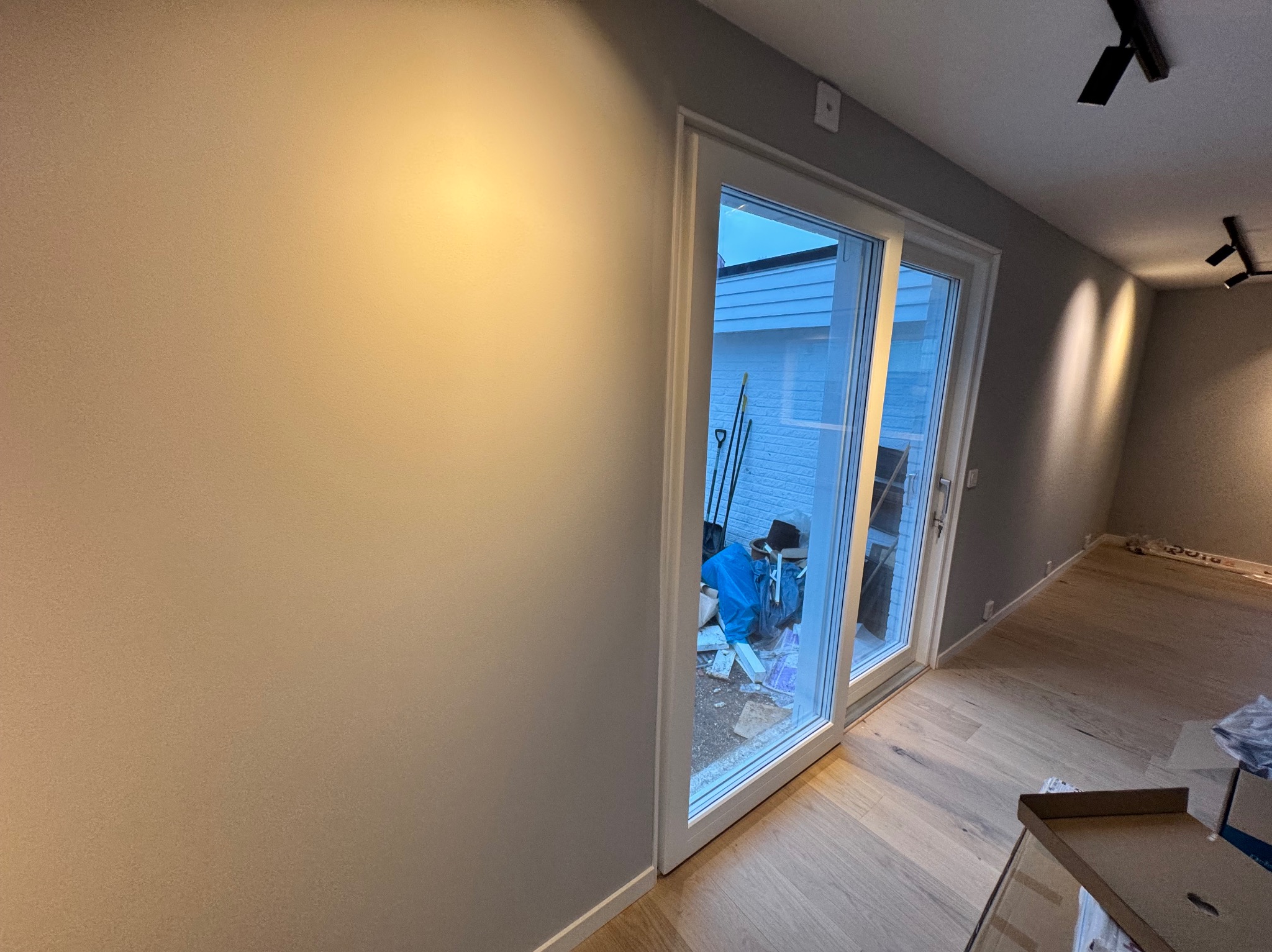 Świeżo pomalowane ściany w odcieniach szarości, widoczne przesuwne drzwi balkonowe z widokiem na podwórze z narzędziami ogrodniczymi i materiałami budowlanymi, parkiet na podłodze, lampy sufitowe...