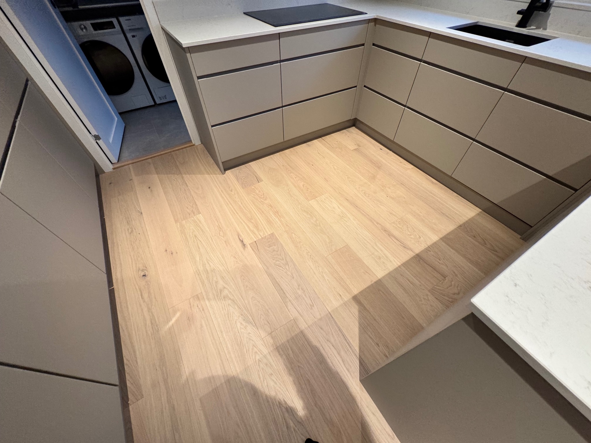 Jasna, drewniana podłoga ułożona w kuchni z szarymi szafkami i białym blatem, widoczne pralki w pomieszczeniu obok.
