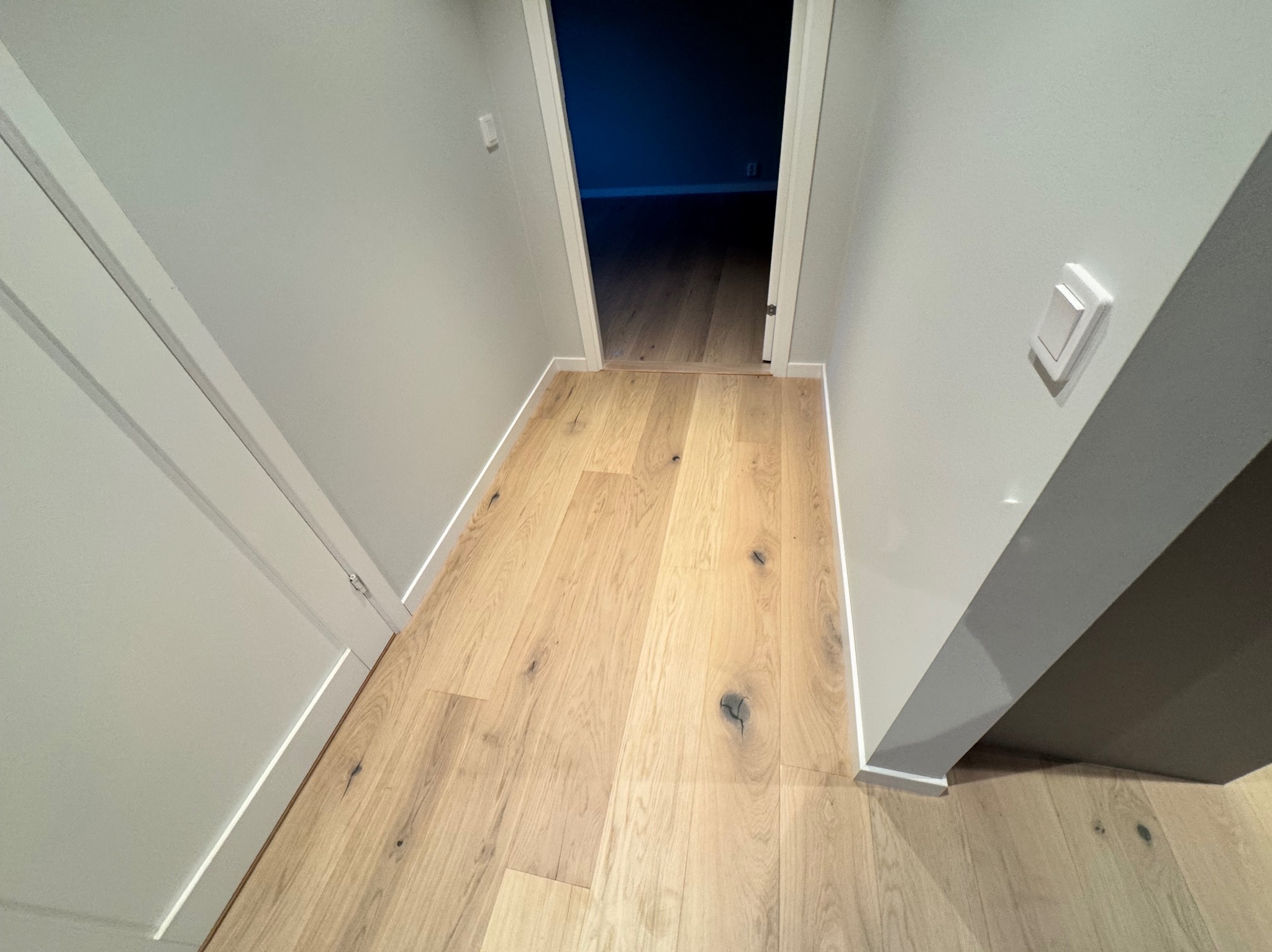 Jasny parkiet z widocznymi sękami w korytarzu z szarymi ścianami i białymi listwami przypodłogowymi, perspektywa w kierunku ciemniejszego pokoju.