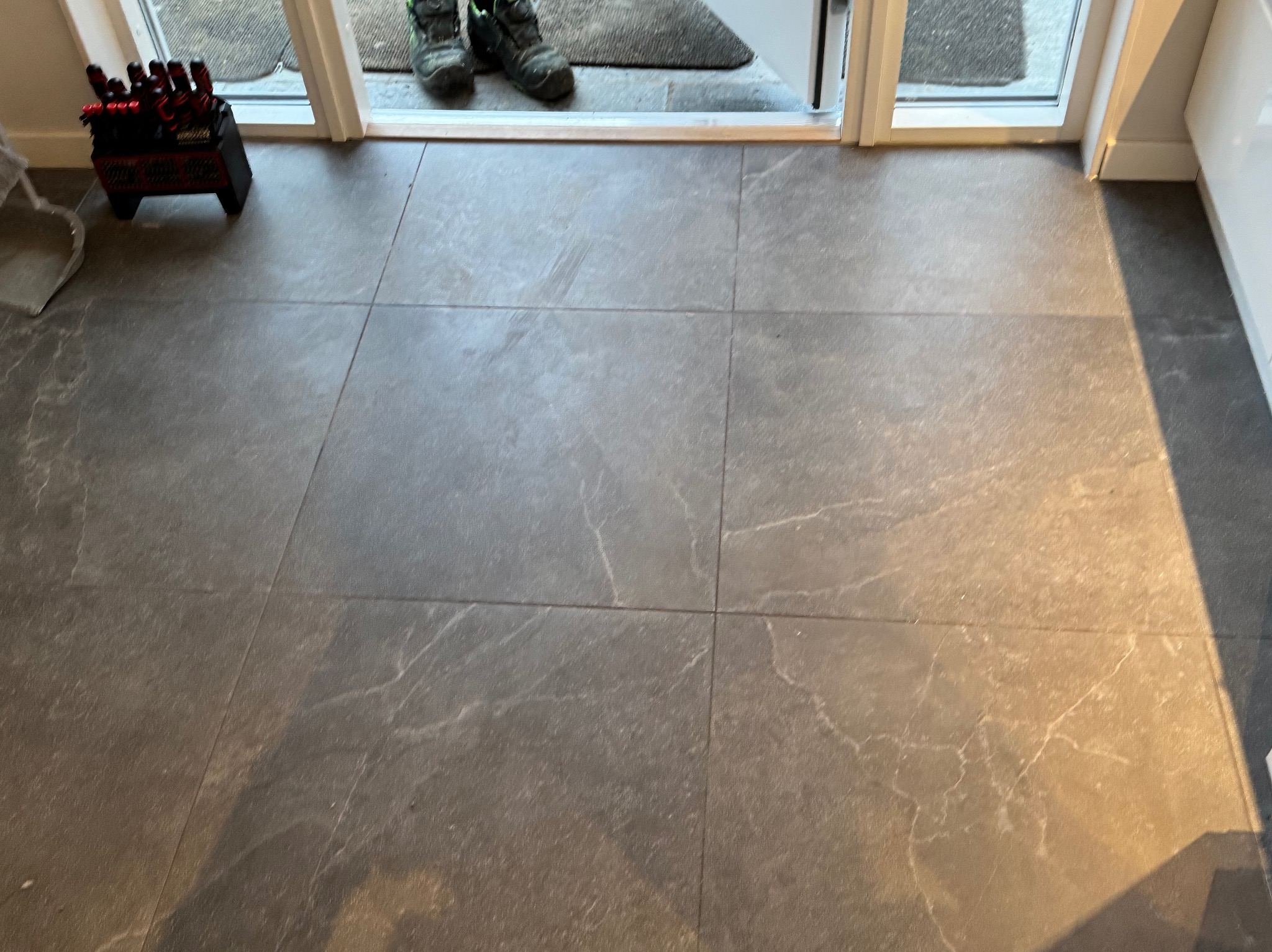Szare, kwadratowe płytki podłogowe z marmurkowym wzorem, widoczne buty robocze przy progu i pojemnik z narzędziami w rogu pomieszczenia.