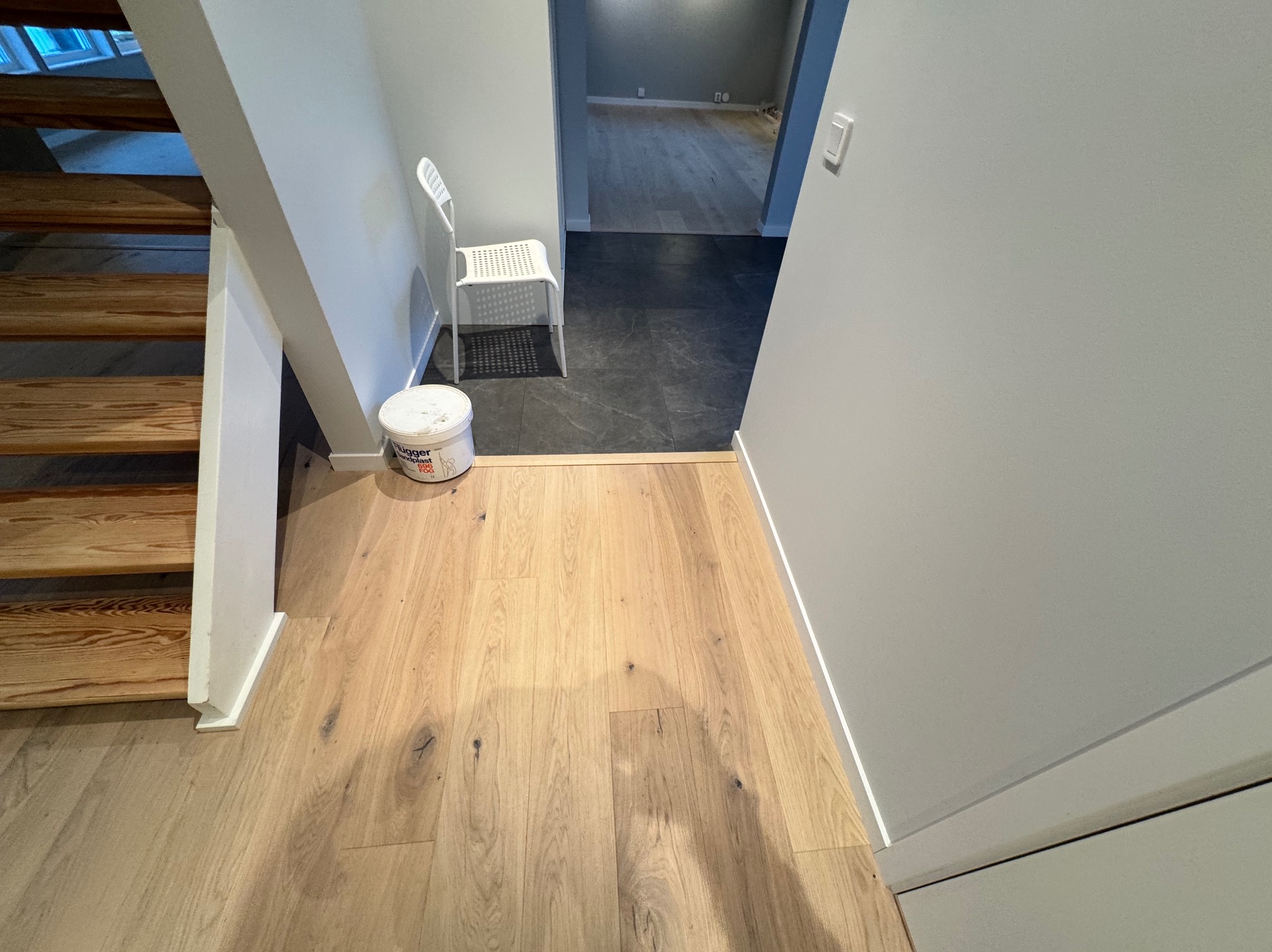 Fragment korytarza z drewnianymi schodami, świeżo położonym parkietem i pomalowanymi na szaro ścianami. Białe krzesło i wiadro farby w tle.