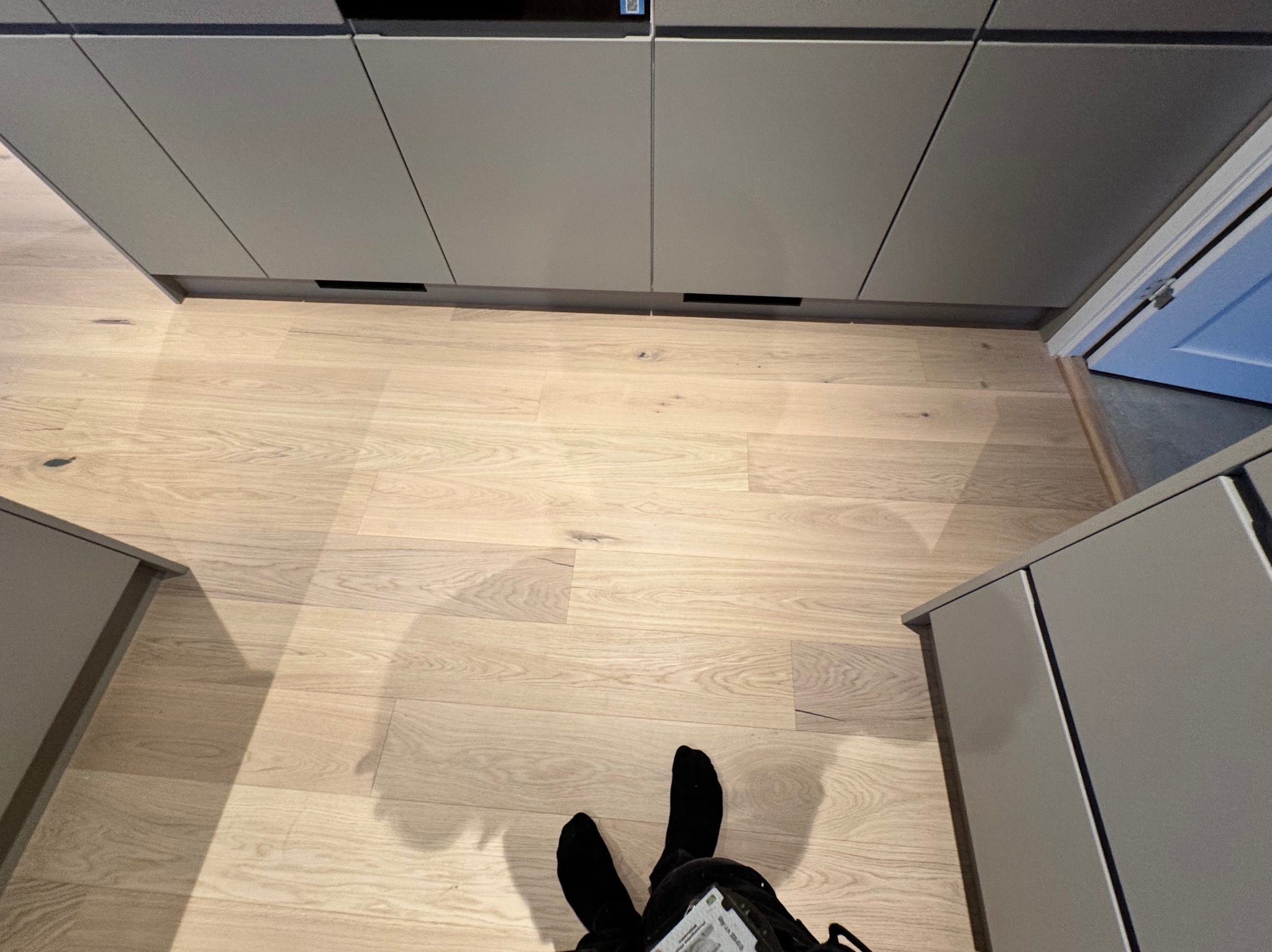Jasny parkiet ułożony w jodełkę francuską, widok z góry na stopy w czarnych skarpetach i fragment szafek kuchennych w kolorze szarym.