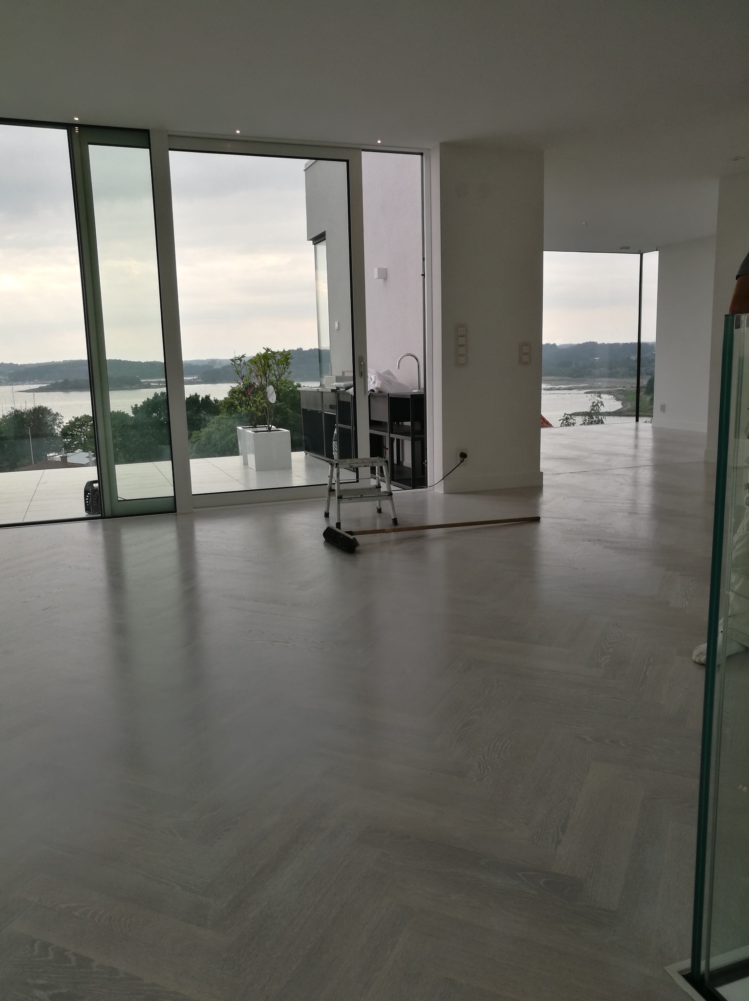 Jasnoszara podłoga ułożona w jodełkę w przestronnym, minimalistycznym wnętrzu z widokiem na jezioro przez duże okna; drabina i miotła sugerują prace wykończeniowe.