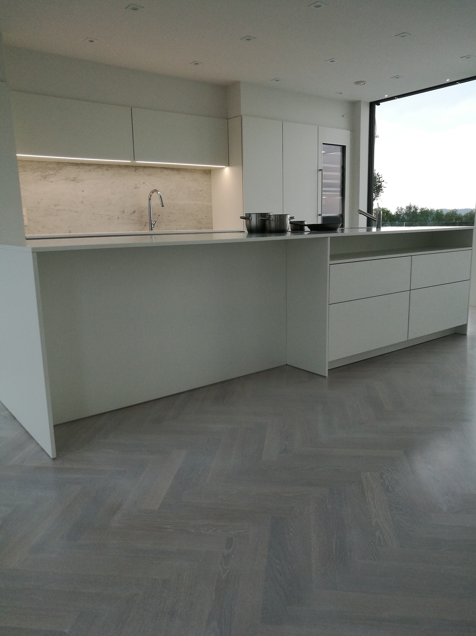 Nowoczesna kuchnia z białą zabudową, wyspą i parkietem ułożonym we wzór jodełki francuskiej, widok na zewnątrz przez duże okno.