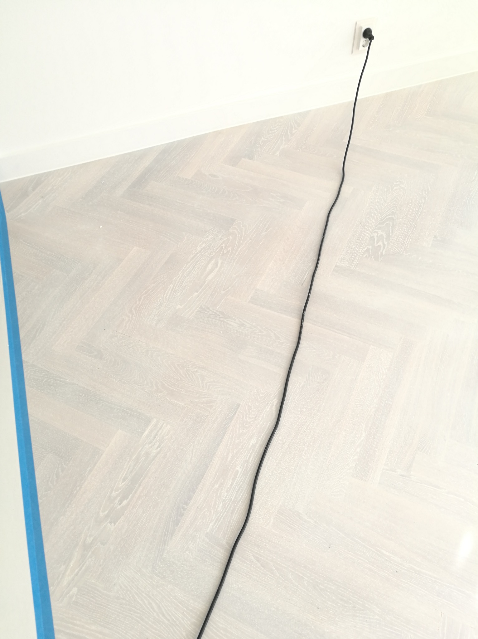 Jasny parkiet ułożony we wzór jodełki, zabezpieczony niebieską taśmą malarską przy ścianie, widoczny kabel zasilający wpięty do gniazdka.