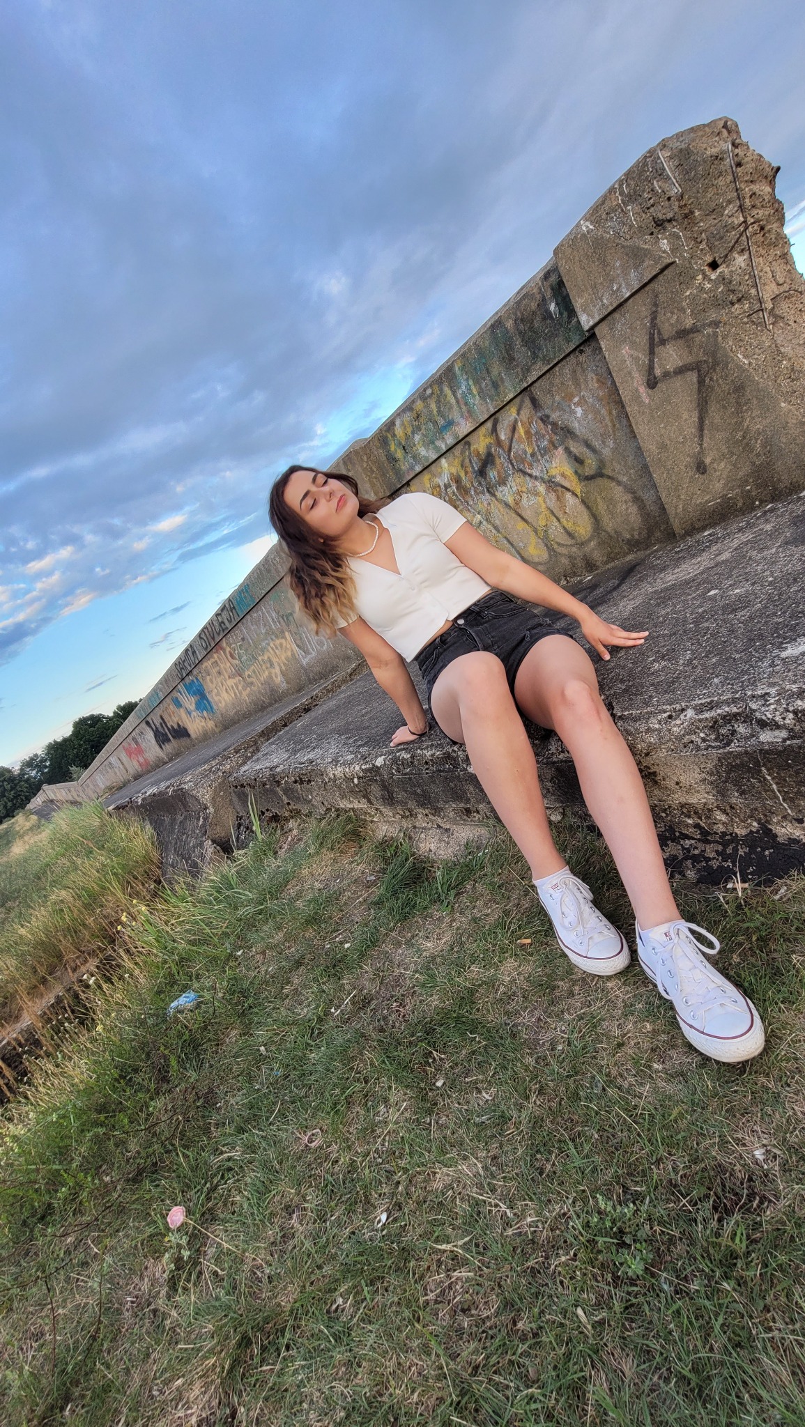 Młoda kobieta w białej bluzce i czarnych szortach pozuje siedząc na betonowym murze z graffiti, z zamkniętymi oczami i rozluźnioną postawą, na tle pochmurnego nieba i trawy.