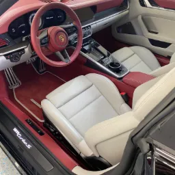 Wnętrze luksusowego samochodu Porsche 911 Turbo S z jasną skórzaną tapicerką i czerwonymi akcentami, widok z otwartych drzwi kierowcy, ukazujący deskę rozdzielczą, kierownicę z logo Porsche i fotele.