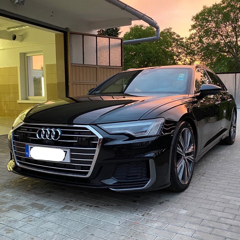 Błyszczący, czarny samochód osobowy Audi A6 Avant po umyciu, zaparkowany na kostce brukowej przed garażem w promieniach zachodzącego słońca.