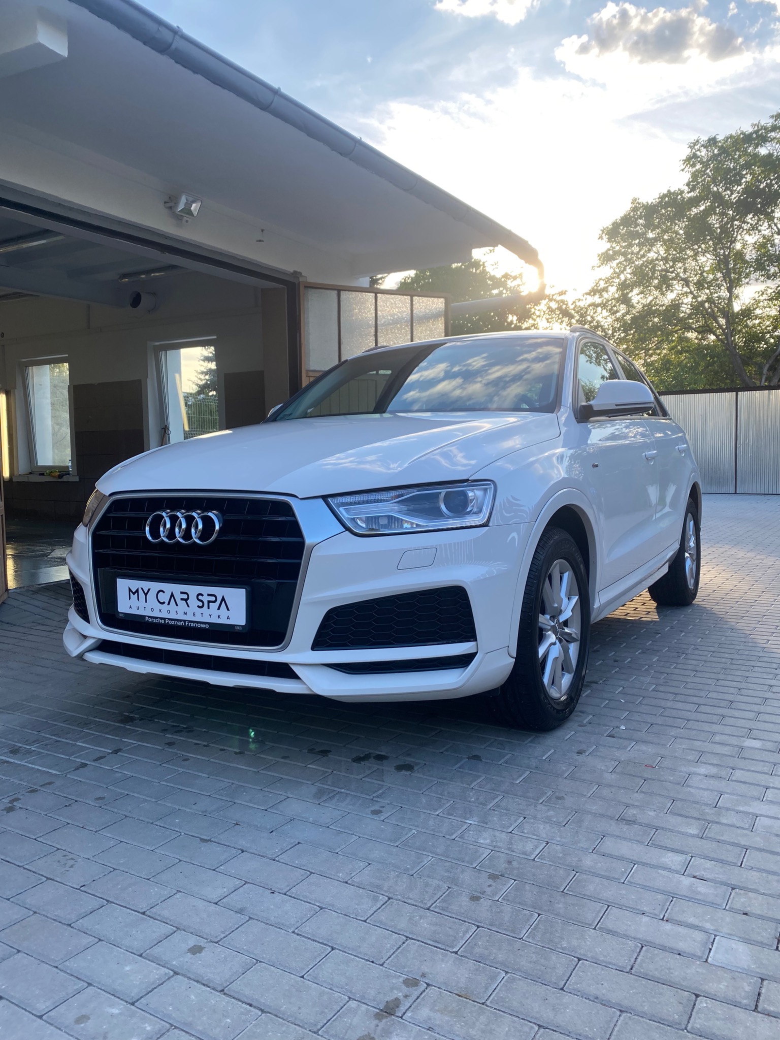 Białe Audi Q3 po zabiegach pielęgnacyjnych, zaparkowane przed myjnią z otwartą bramą w słoneczny dzień.