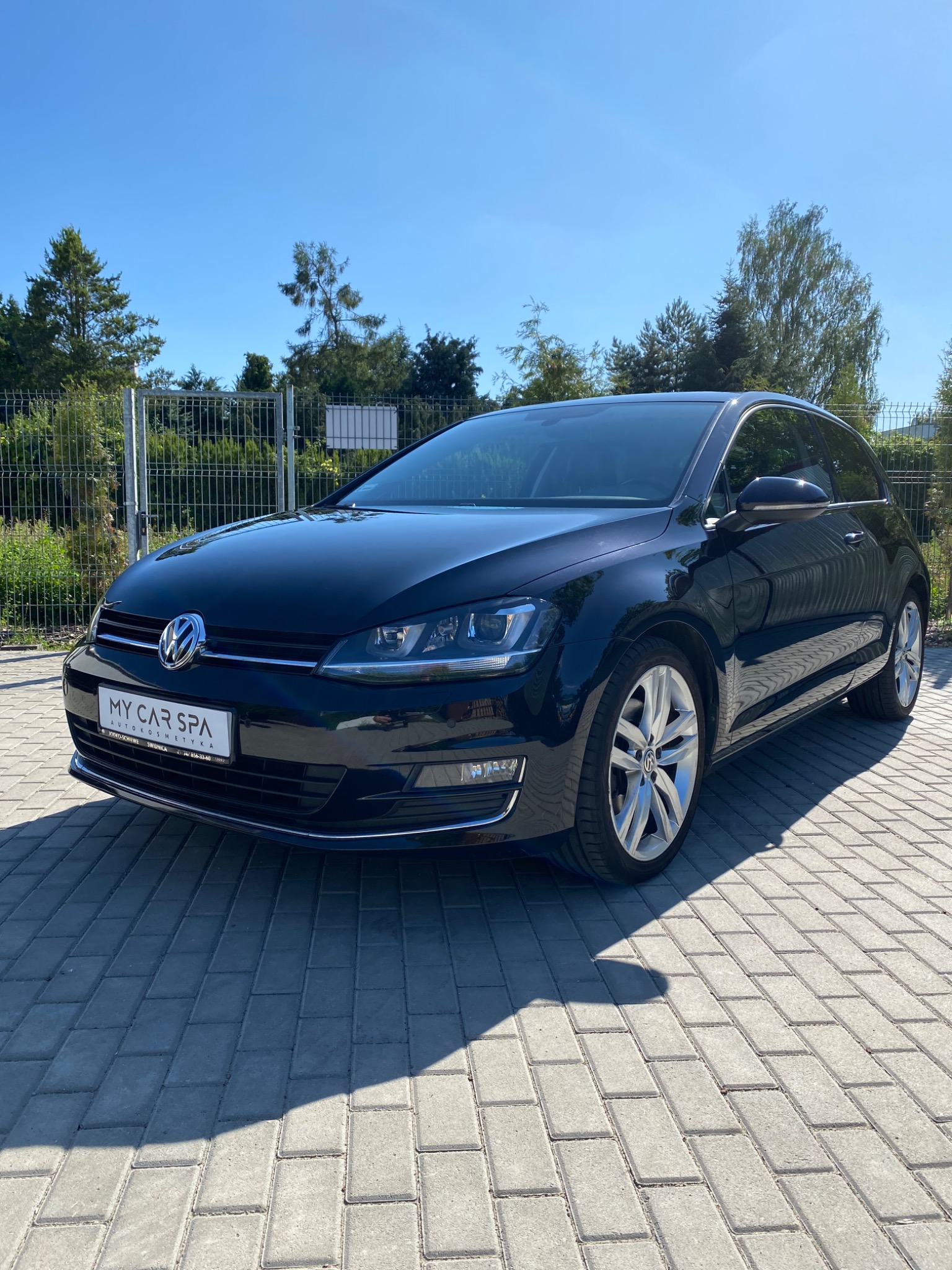 Błyszczący, czarny Volkswagen Golf po wizycie w myjni, zaparkowany na szarym bruku w słoneczny dzień. Na tablicy rejestracyjnej widoczne logo 'My Car Spa'.