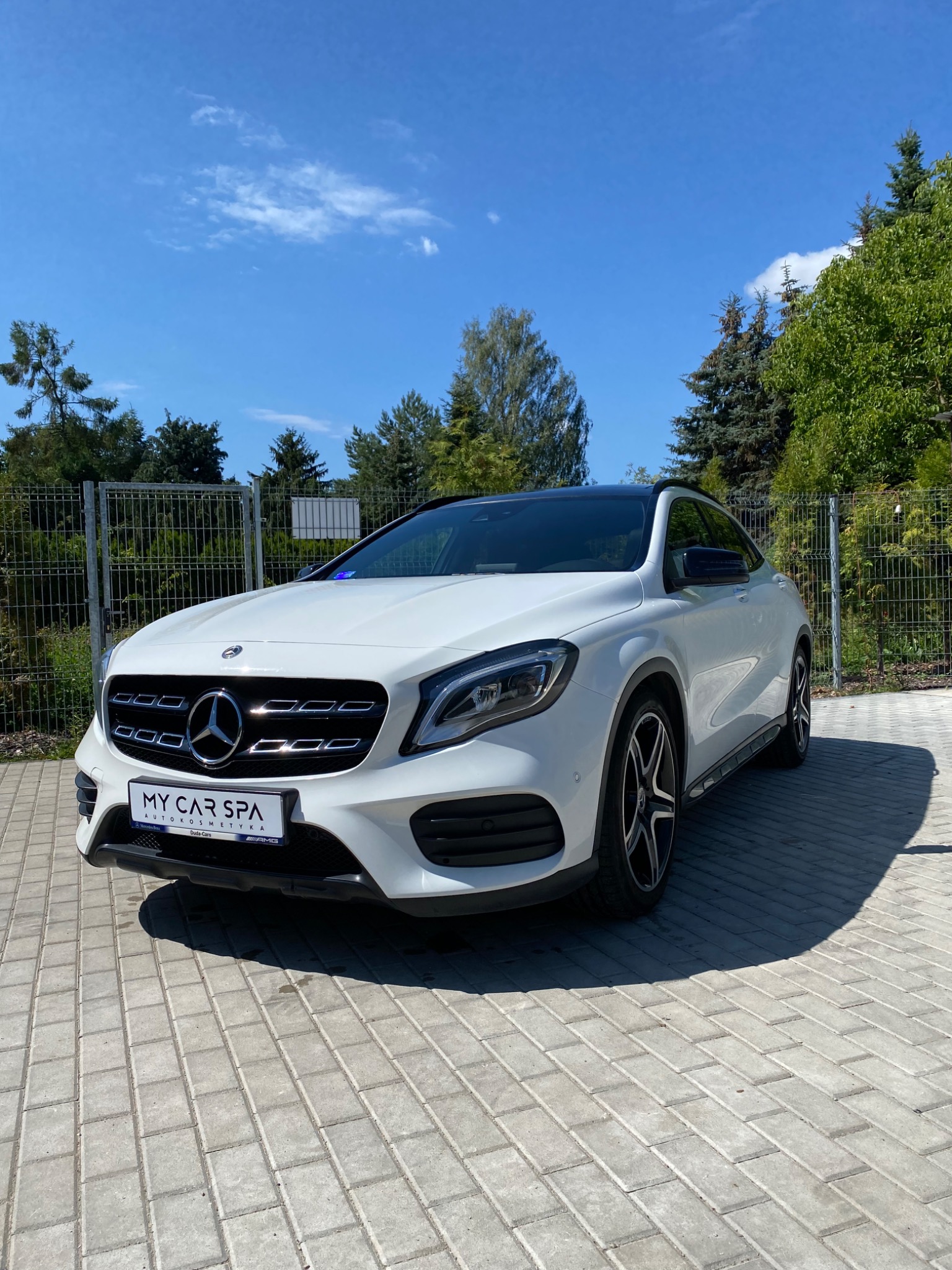 Biały Mercedes-Benz GLA po usłudze auto detailingu, zaparkowany na kostce brukowej przed ogrodzeniem, z widocznym logo My Car Spa na tablicy rejestracyjnej.