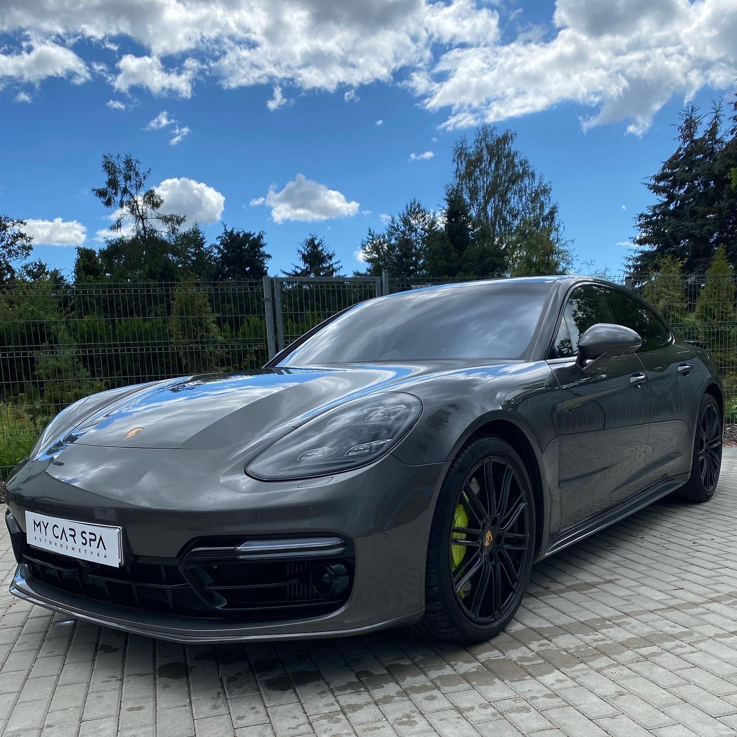 Szary samochód Porsche Panamera po detailingu, z czarnymi felgami i neonowo-zielonymi zaciskami hamulcowymi, zaparkowany na kostce brukowej, na tle ogrodzenia i zieleni.