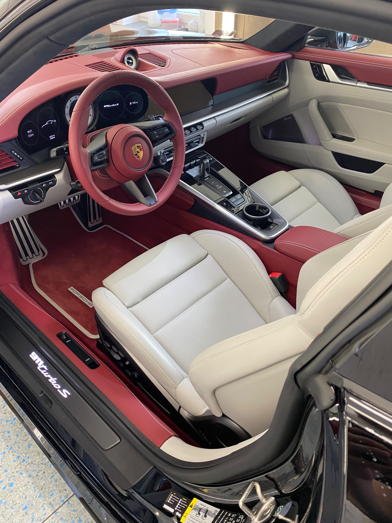 Wnętrze luksusowego samochodu Porsche 911 Turbo S z jasną skórzaną tapicerką i czerwonymi akcentami, widok z otwartych drzwi kierowcy, ukazujący deskę rozdzielczą, kierownicę z logo Porsche i fotele.