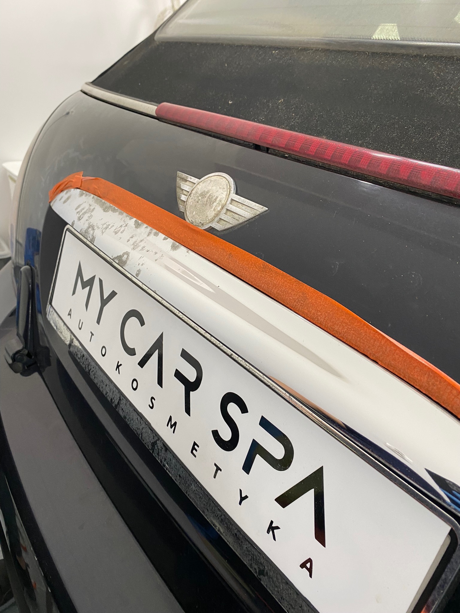 Zabrudzony tył czarnego samochodu Mini Cooper z naklejoną pomarańczową taśmą malarską zabezpieczającą chromowaną listwę z napisem 'MY CAR SPA AUTOKOSMETYKA' przed polerowaniem.