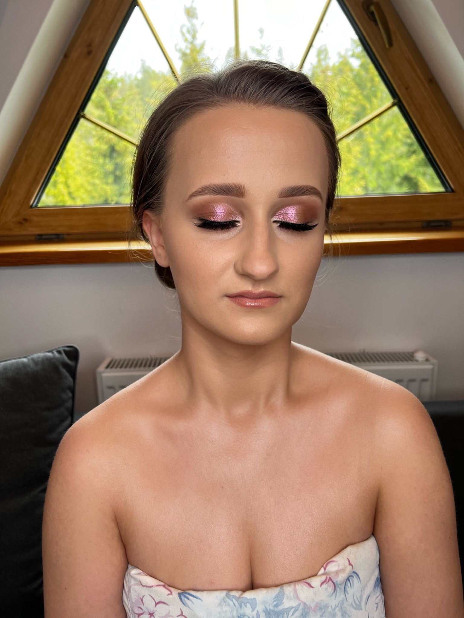 Portret kobiety z zamkniętymi oczami, prezentującej delikatny, różowy makijaż oczu z wydłużoną kreską i błyszczącymi ustami na tle okna w kształcie trójkąta.