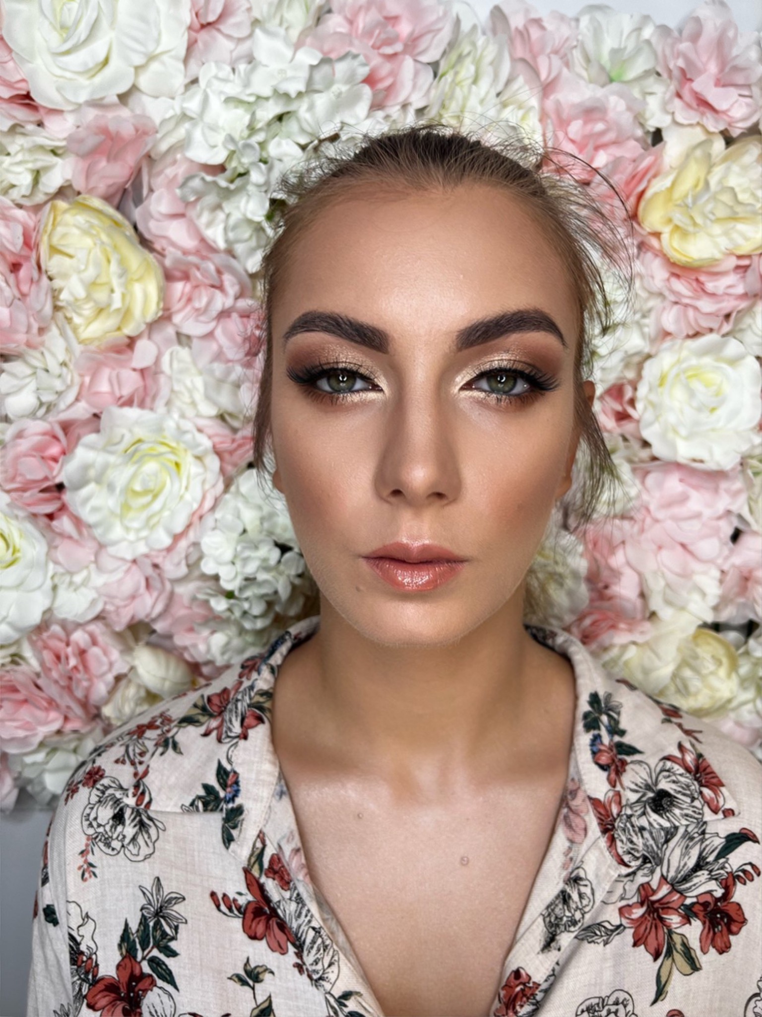 Portret kobiety z profesjonalnym makijażem oczu w odcieniach brązu i złota, błyszczące usta, na tle ściany z różowych i białych kwiatów.