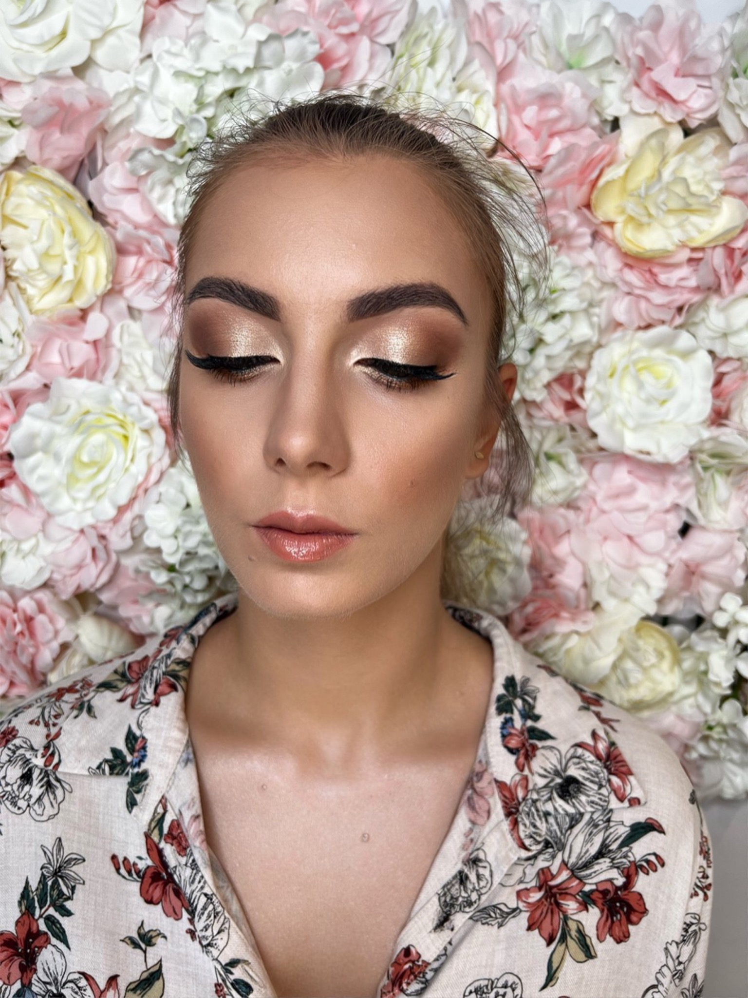 Portret kobiety z zamkniętymi oczami, prezentującej złoty makijaż oczu i błyszczące usta na tle ściany z kwiatów w odcieniach różu i bieli.