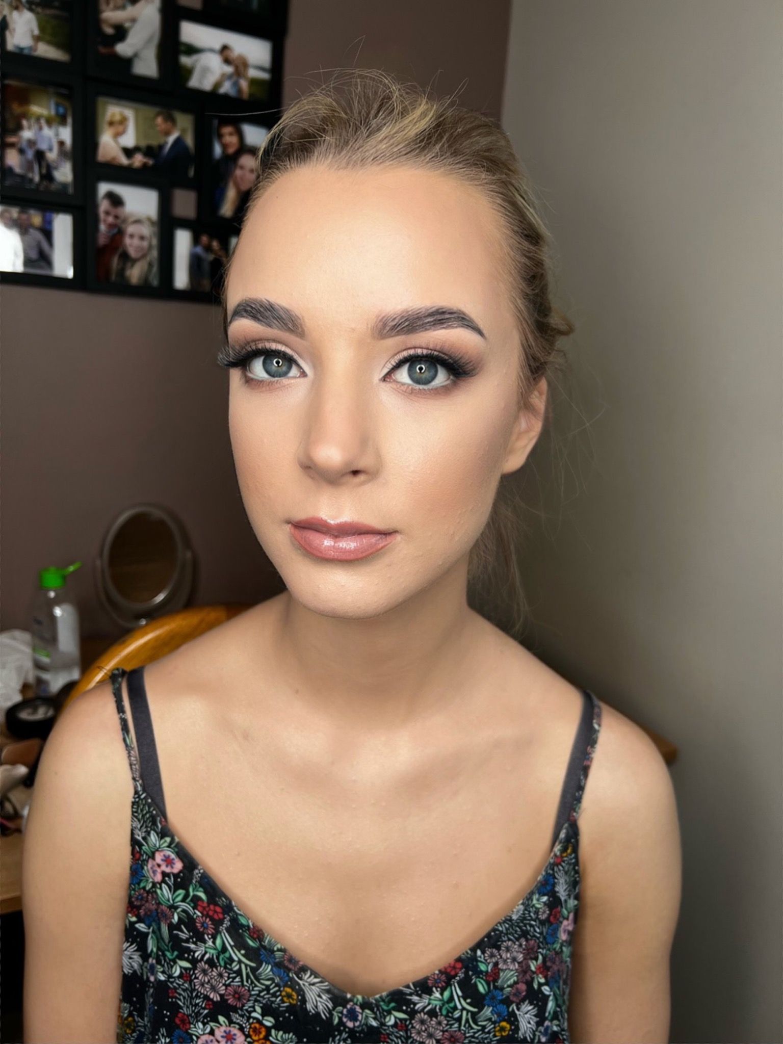 Portret młodej kobiety z delikatnym makijażem, podkreślone oczy z długimi rzęsami, błyszczące usta, w tle ściana z ramkami zdjęć.