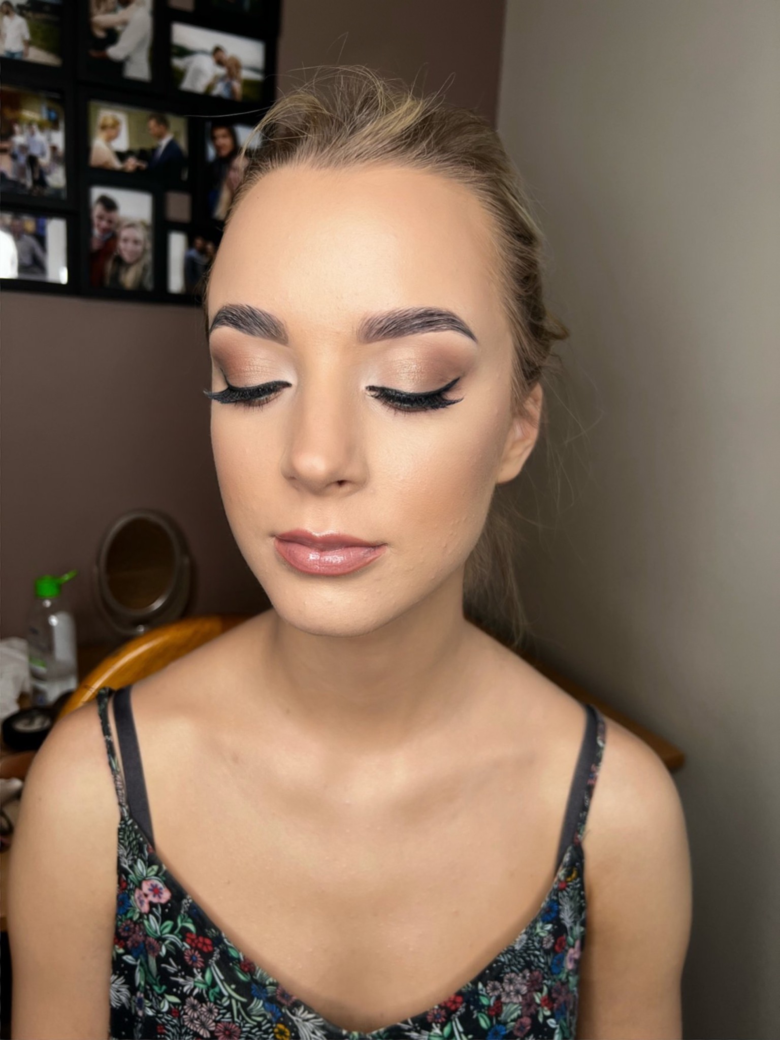 Portret młodej kobiety z zamkniętymi oczami, prezentujący delikatny, brązowy makijaż oczu z wyraźną kreską, podkreślone brwi i błyszczące usta; w tle ramka ze zdjęciami.