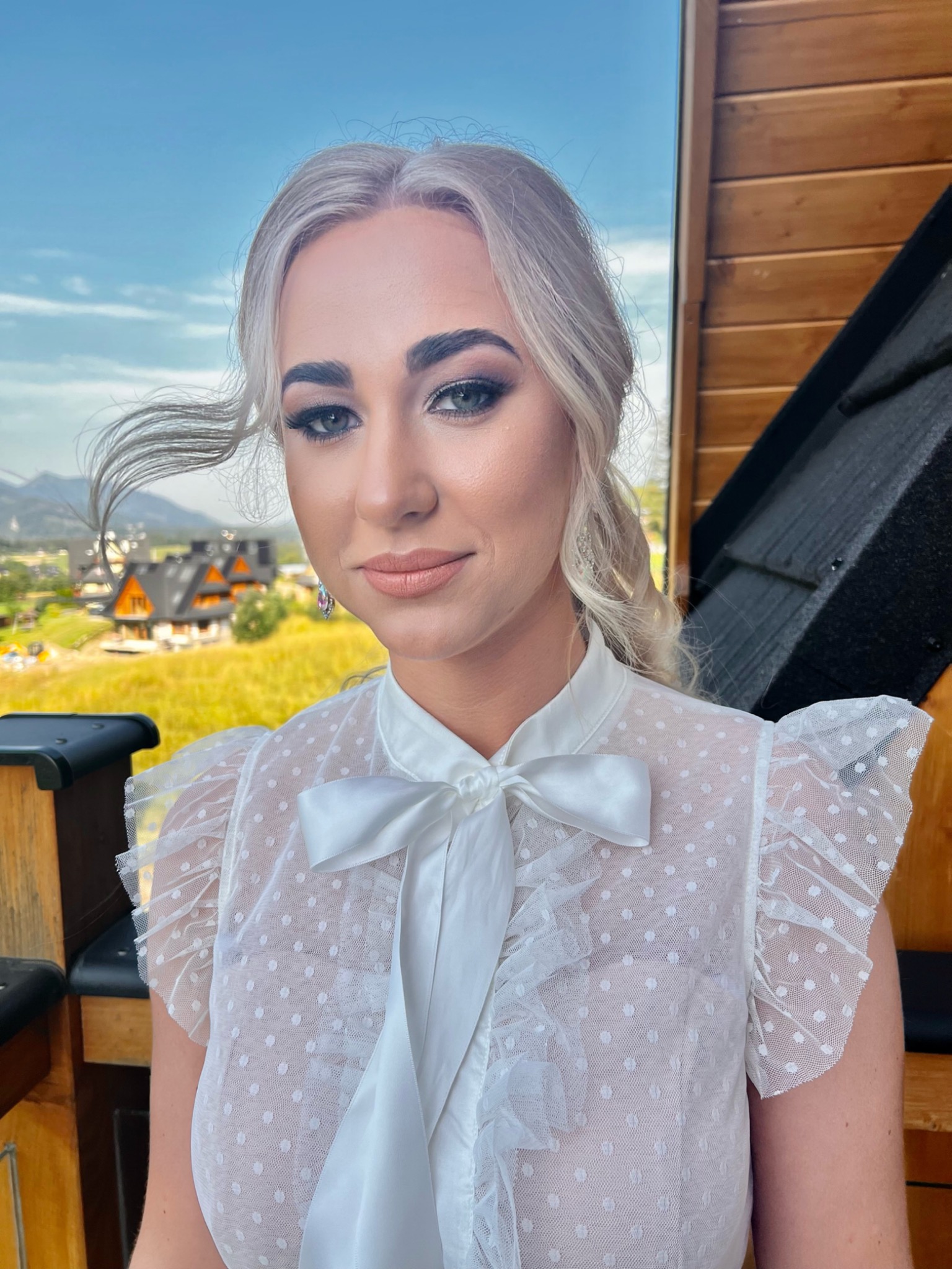 Portret kobiety z profesjonalnym makijażem, w tle górski krajobraz z drewnianymi domami, włosy blond powiewają na wietrze, ubrana w białą bluzkę w kropki z kokardą.