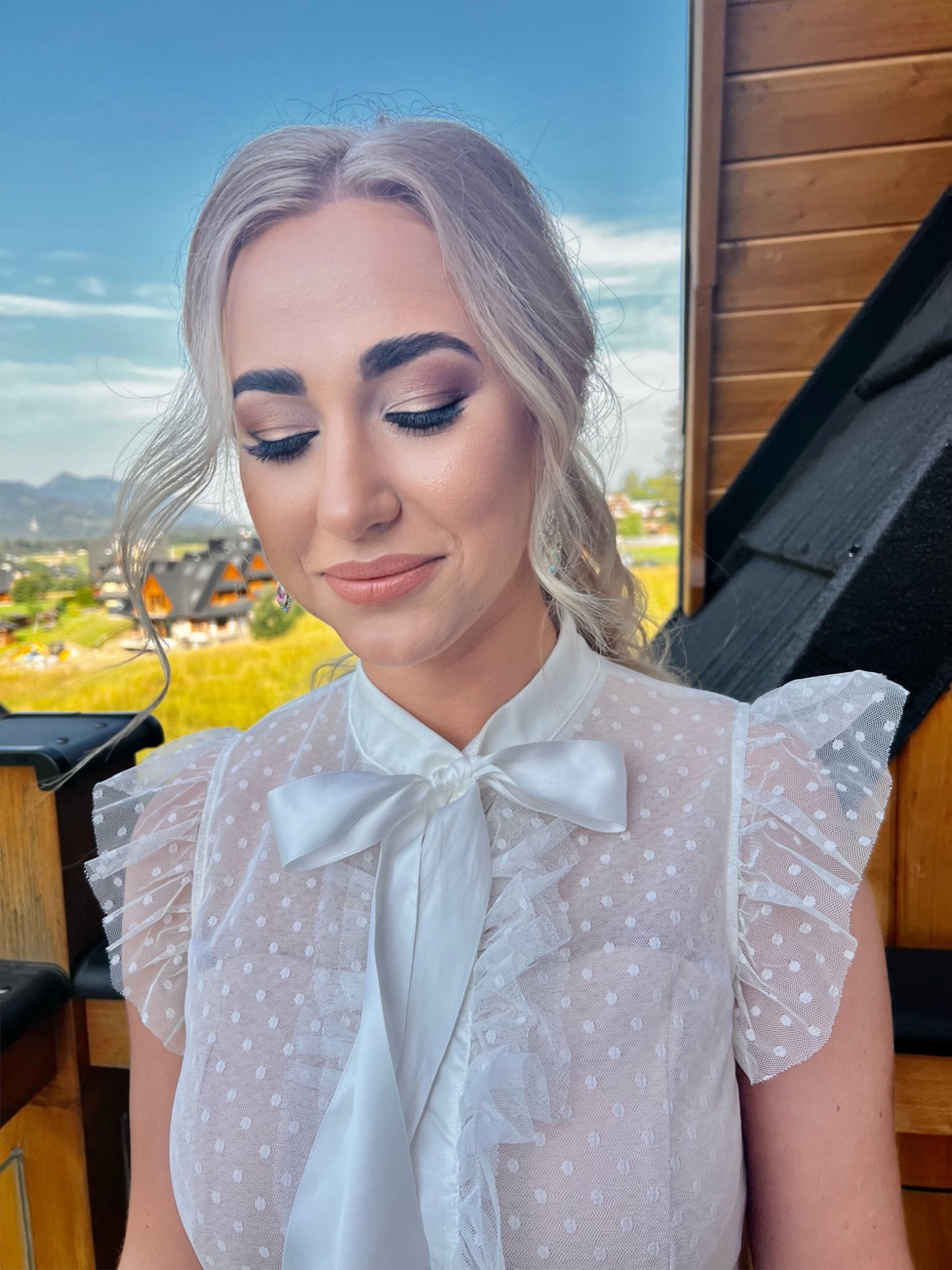 Portret kobiety z delikatnym makijażem oczu w odcieniach brązu i beżu, podkreślonymi rzęsami, oraz subtelnym makijażem ust, na tle górskiego krajobrazu i drewnianej architektury.