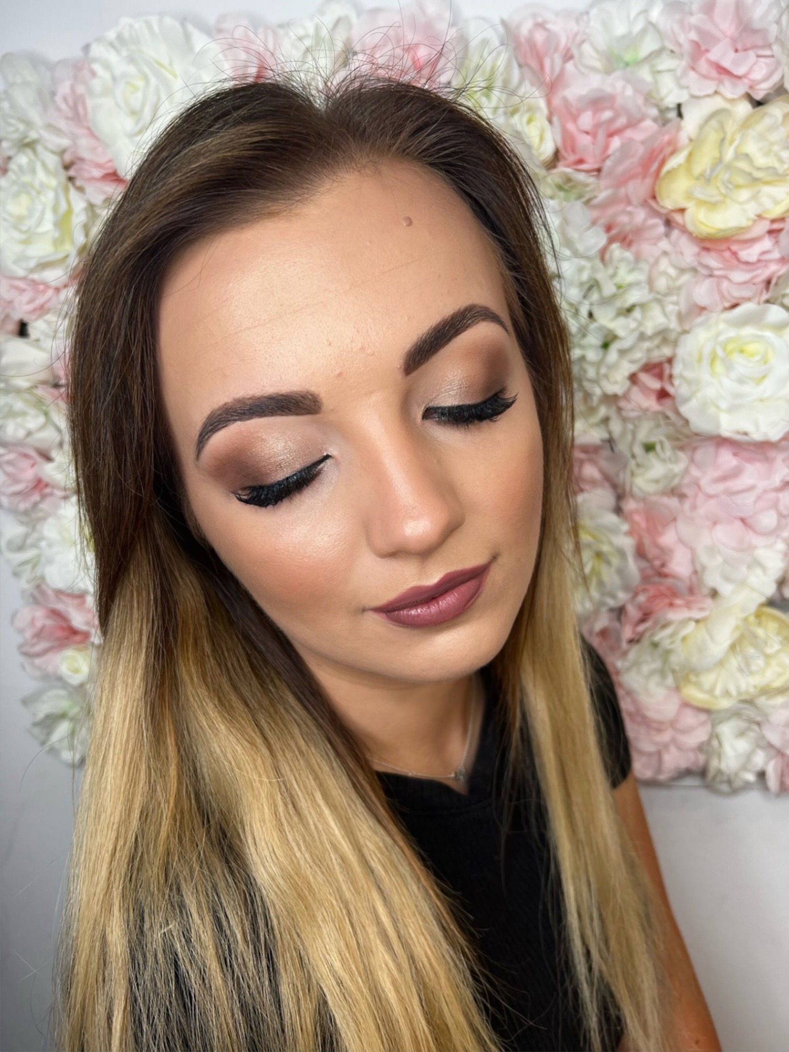 Portret kobiety z zamkniętymi oczami prezentującej profesjonalny, wieczorowy makijaż w odcieniach brązu i złota, z wyraźnie podkreślonymi rzęsami i matowymi ustami w kolorze mauve, na tle ściany...